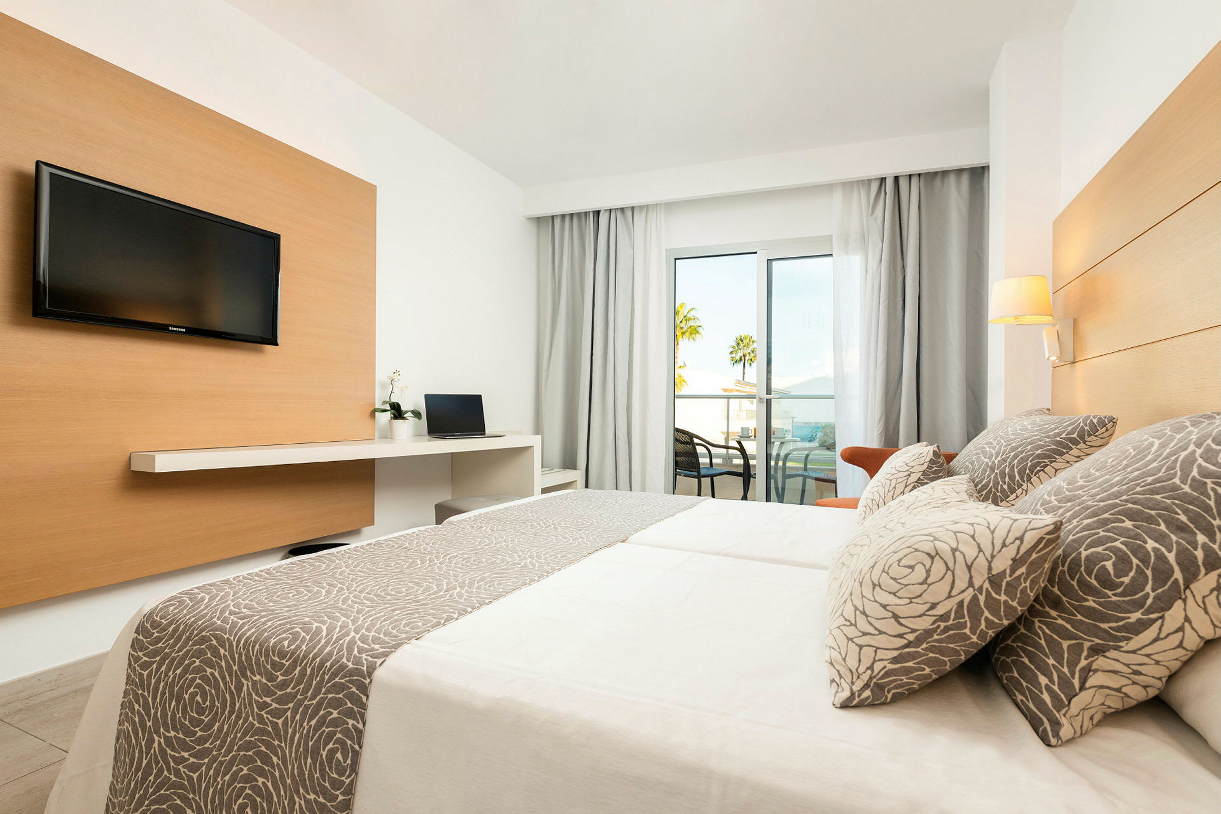 Værelset har et TV, skrivebord med bærbar computer og en seng med mønstrede puder. En balkon med udsigt ses i baggrunden. (AI-genereret tekst)