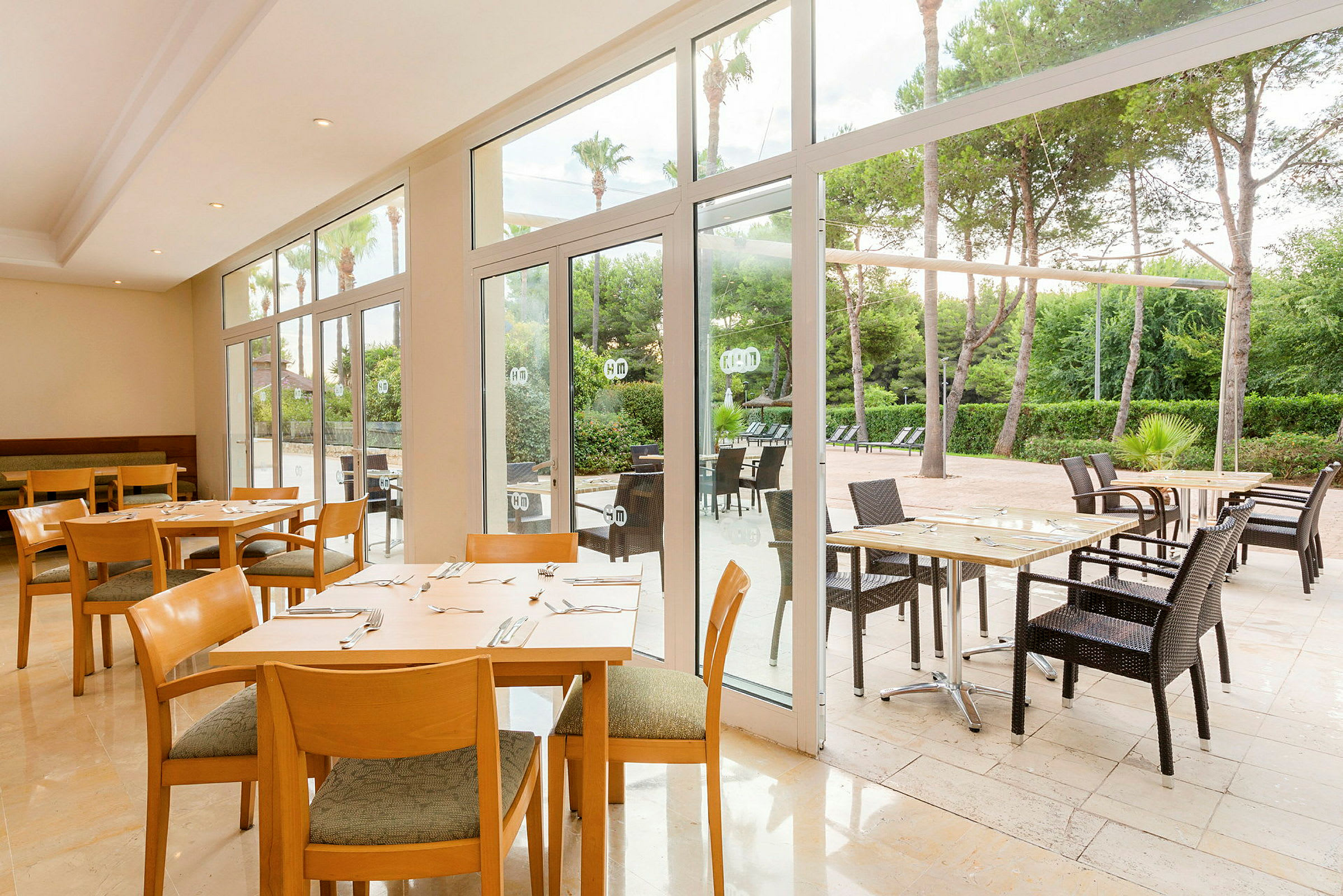 Restaurant med borde og stole både indendørs og udendørs, med udsigt til have og træer. (AI-genereret tekst)