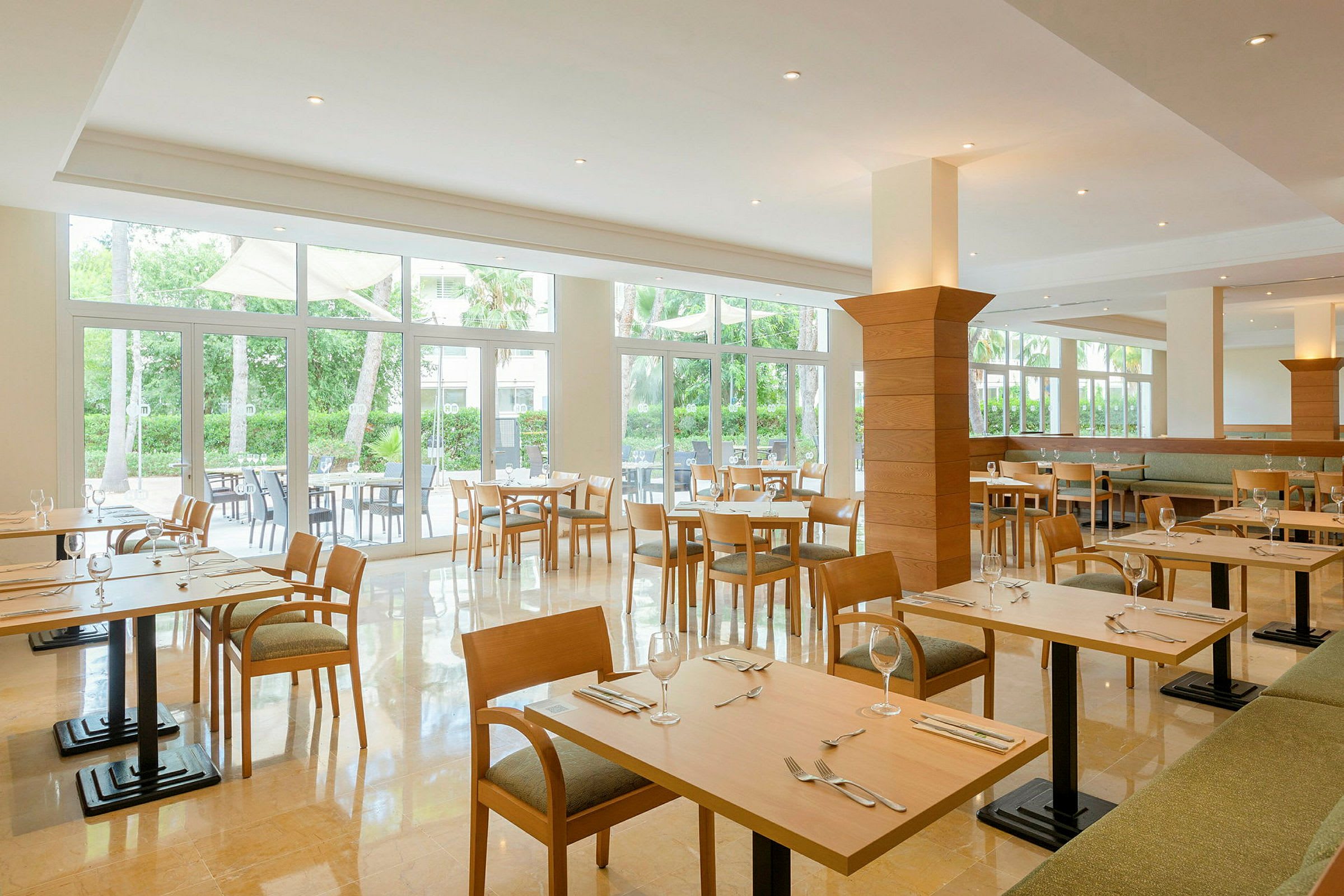 Lyst og rummeligt restaurantområde med træborde og stole, omgivet af store vinduer med udsigt til grønne områder. (AI-genereret tekst)