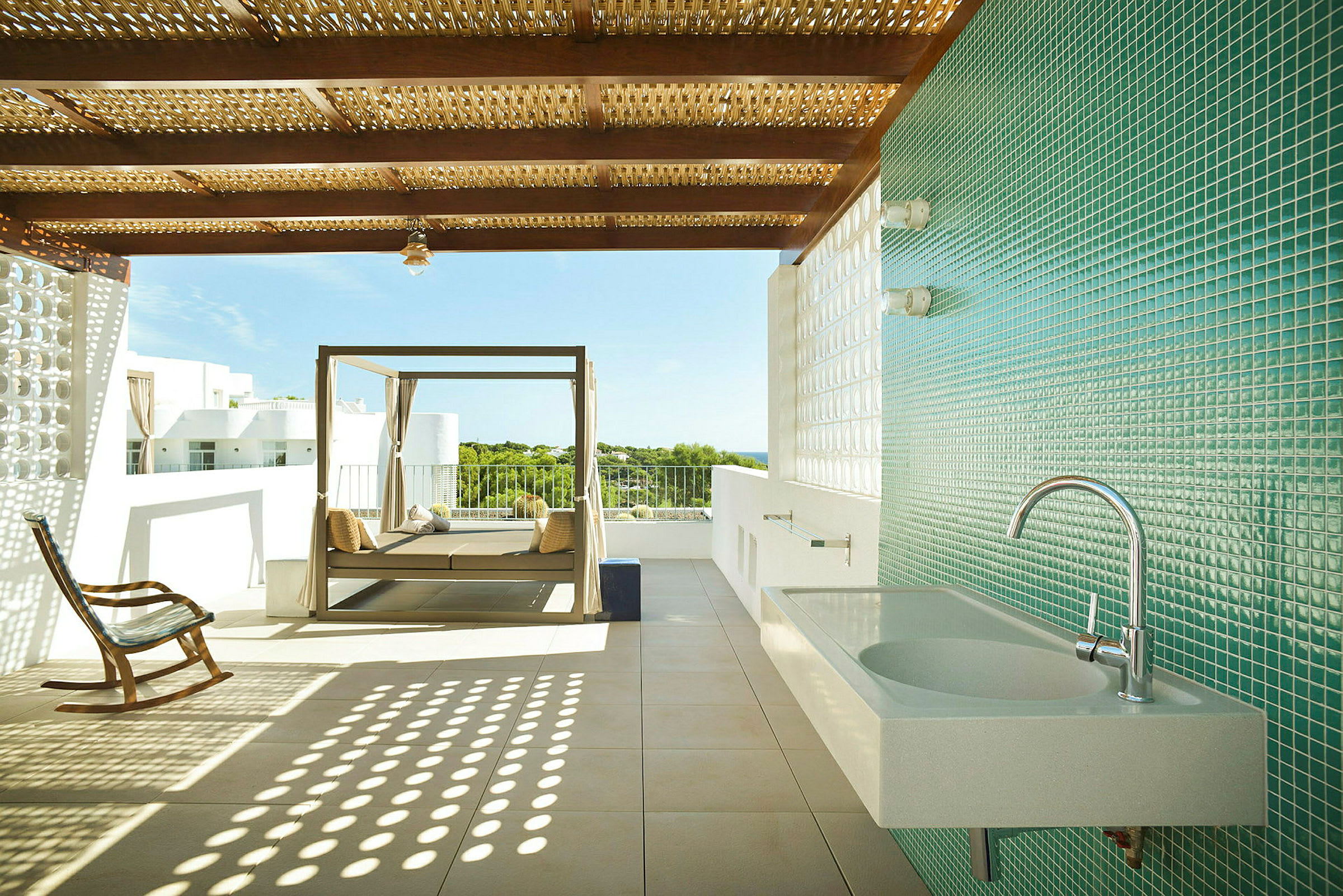 En udendørs terrasse med tag, solseng og en gyngestol samt en væg med mosaik og en vask på den ene side. (AI-genereret tekst)
