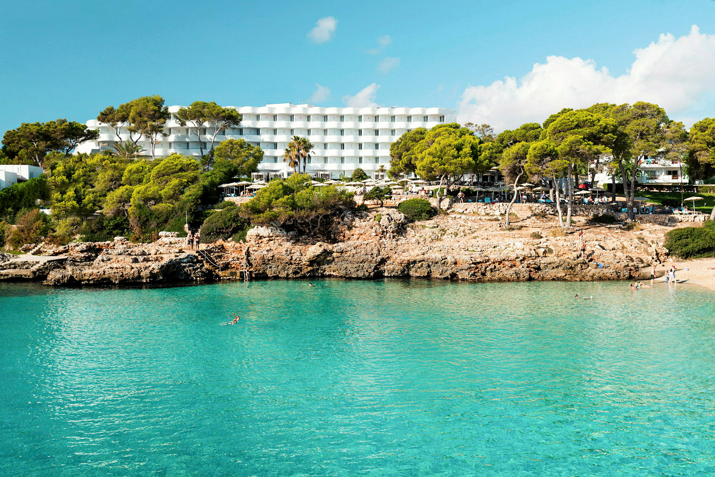 Hotel med hvide bygninger bag en klippefyldt strand med klart blåt vand. (AI-genereret tekst)