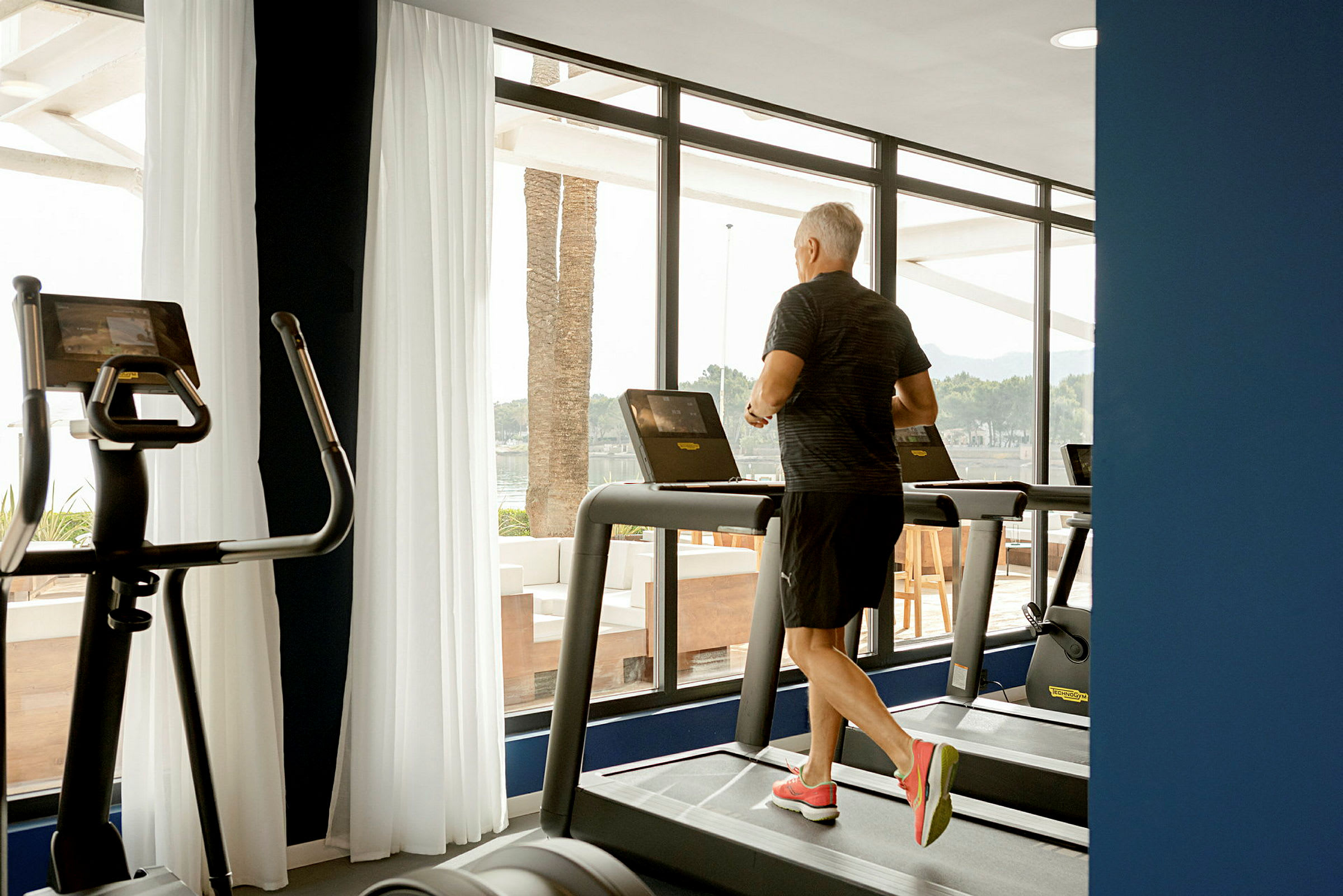 En person løber på et løbebånd i et fitnesscenter med store vinduer. (AI-genereret tekst)