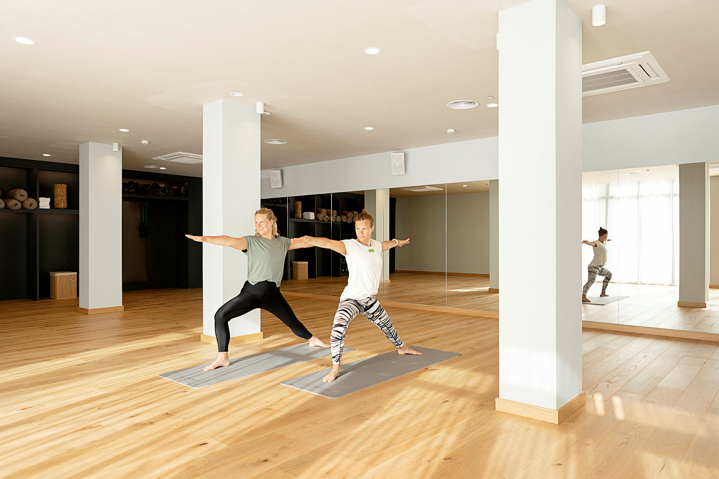 To personer udfører yoga i en lys studie med trægulv og spejlvæg. (AI-genereret tekst)