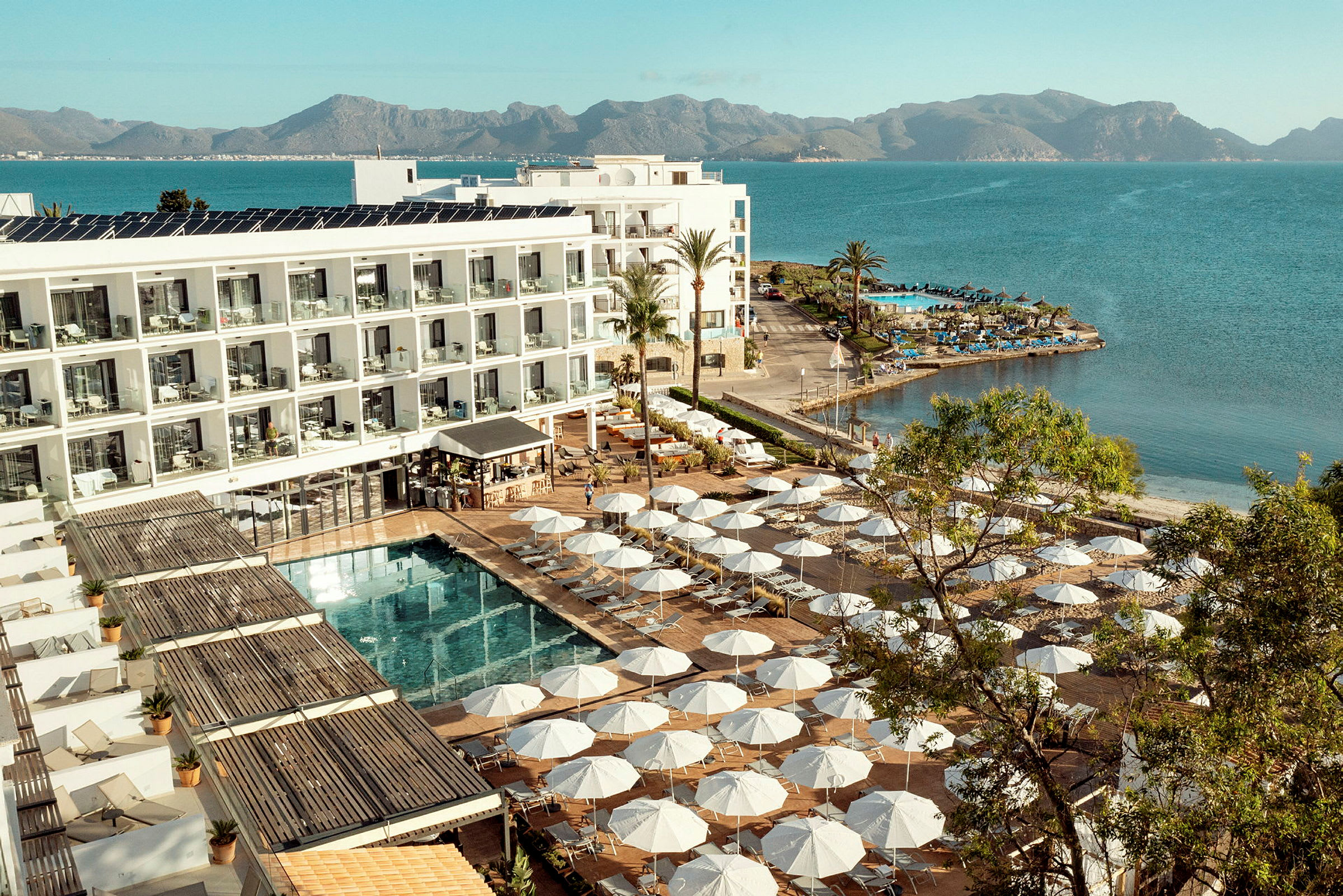 Hotelkompleks ved vandkanten med solsenge og parasoller omkring en pool. (AI-genereret tekst)