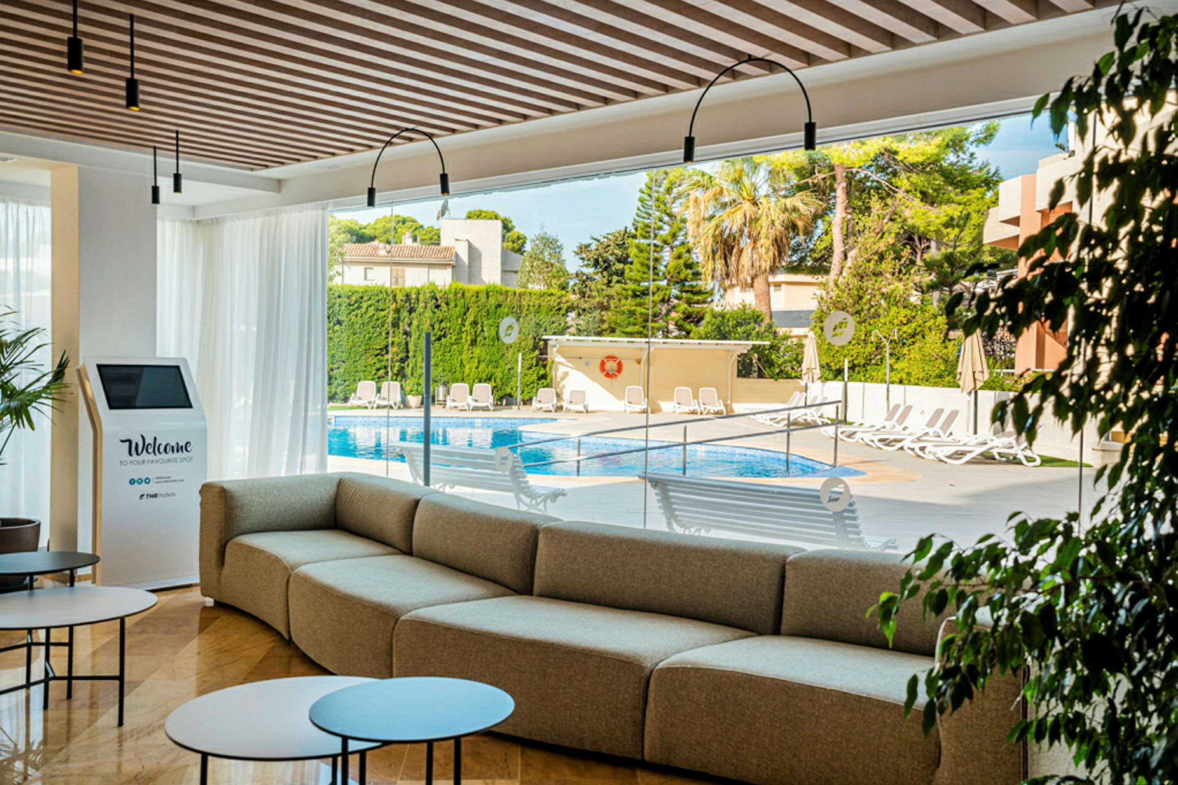 En moderne lounge med sofa, borde og udsigt til poolområdet udenfor glasvæggene. (AI-genereret tekst)