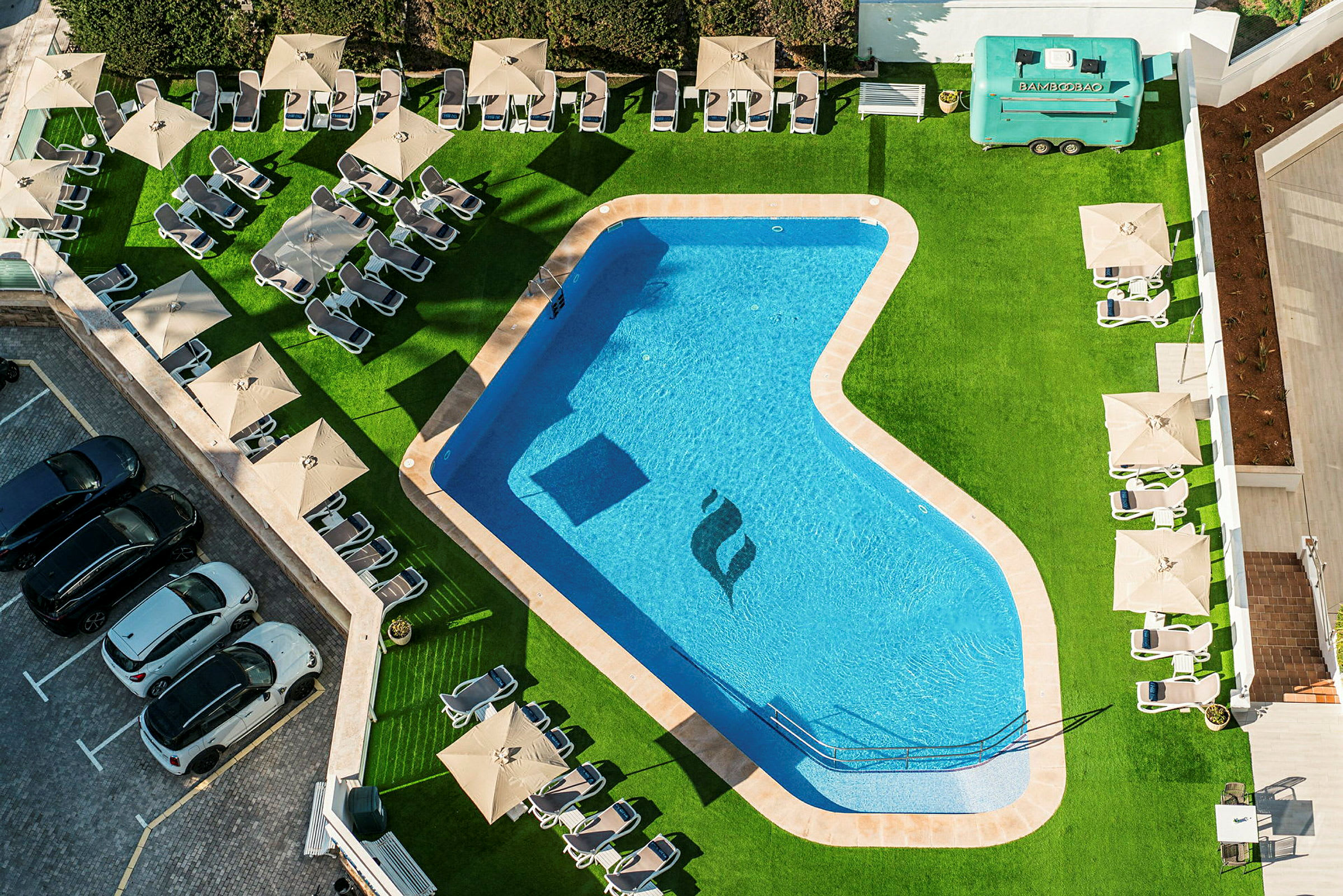 Luftfoto af en udendørs pool omgivet af liggestole og parasoller på græs, med biler parkeret ved siden af. (AI-genereret tekst)