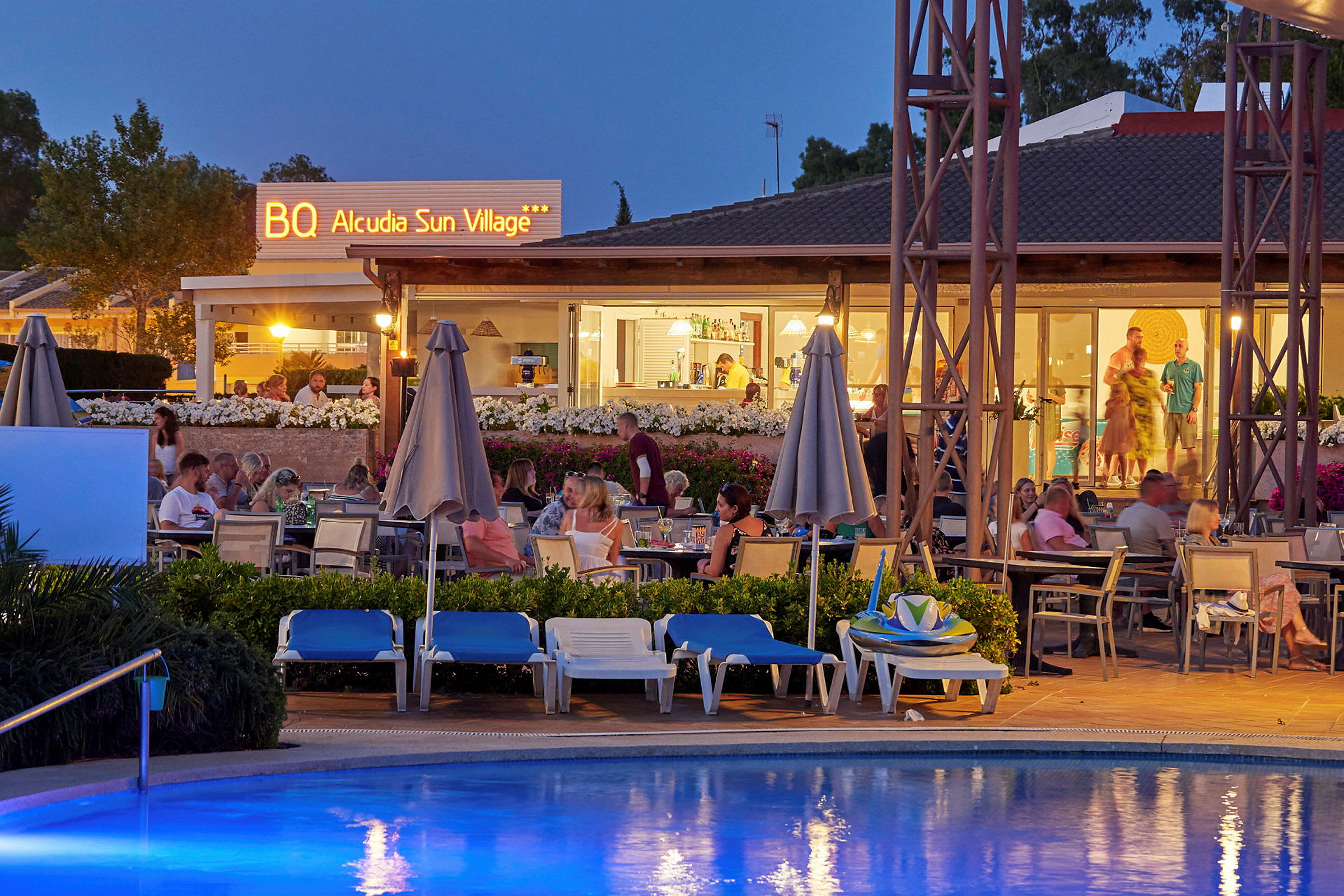 Udendørs serveringsområde ved et hotel med gæster og en pool i forgrunden, om aftenen. (AI-genereret tekst)