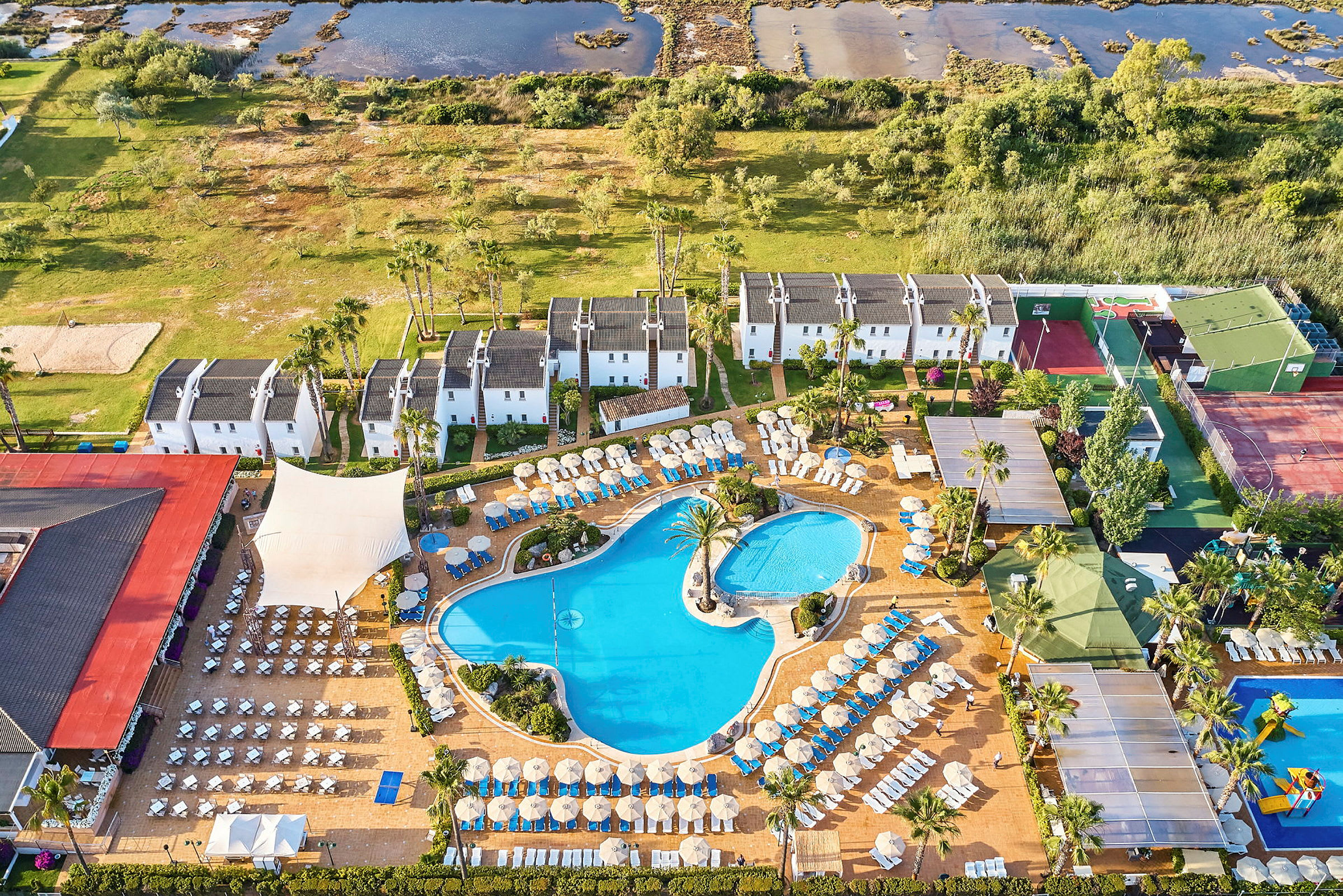 Luftfoto af hotelområde med pools, liggestole og hvidmalede bygninger omgivet af græsarealer. (AI-genereret tekst)