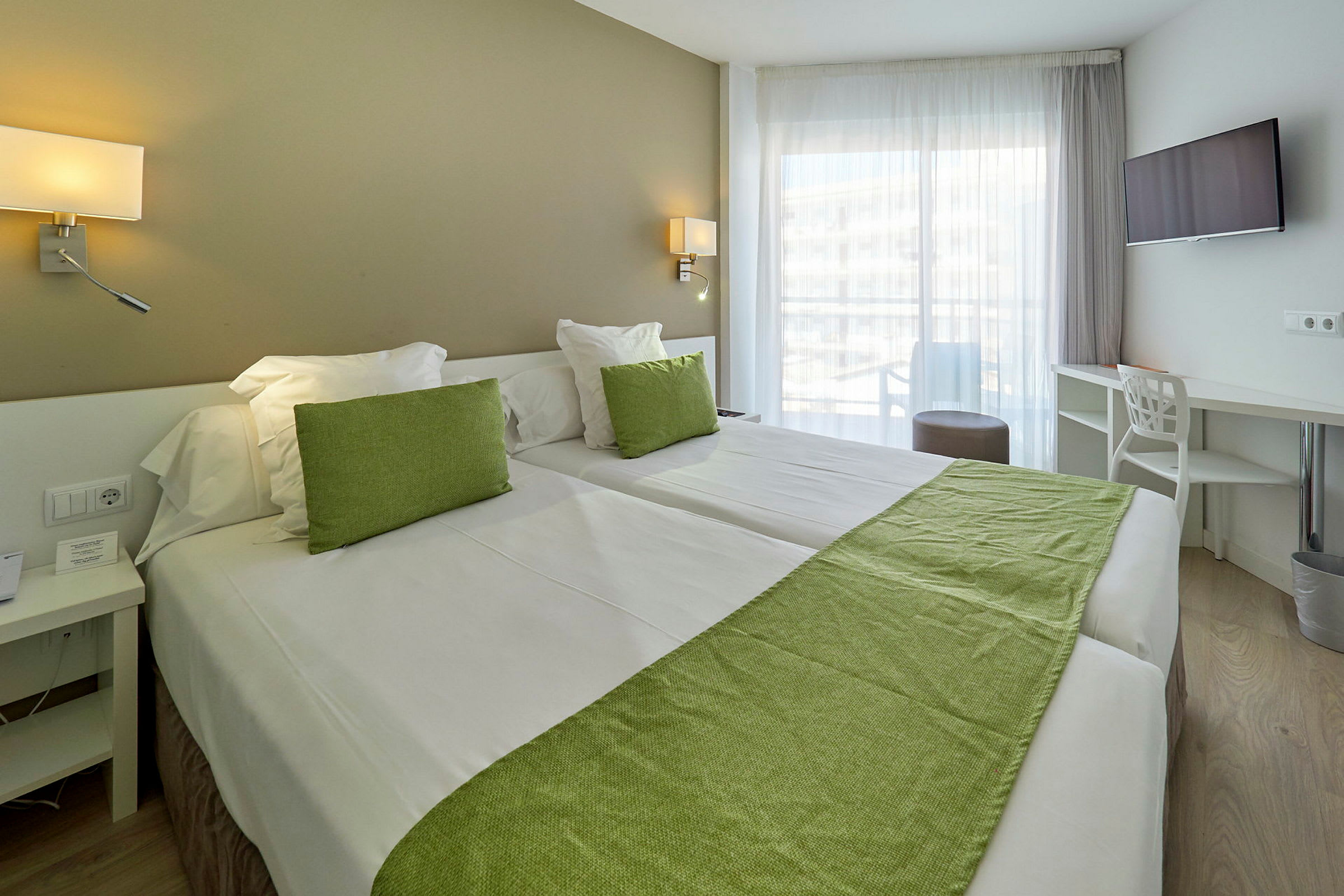 Hotelværelse med to senge, grøn dekoration og store vinduer. (AI-genereret tekst)