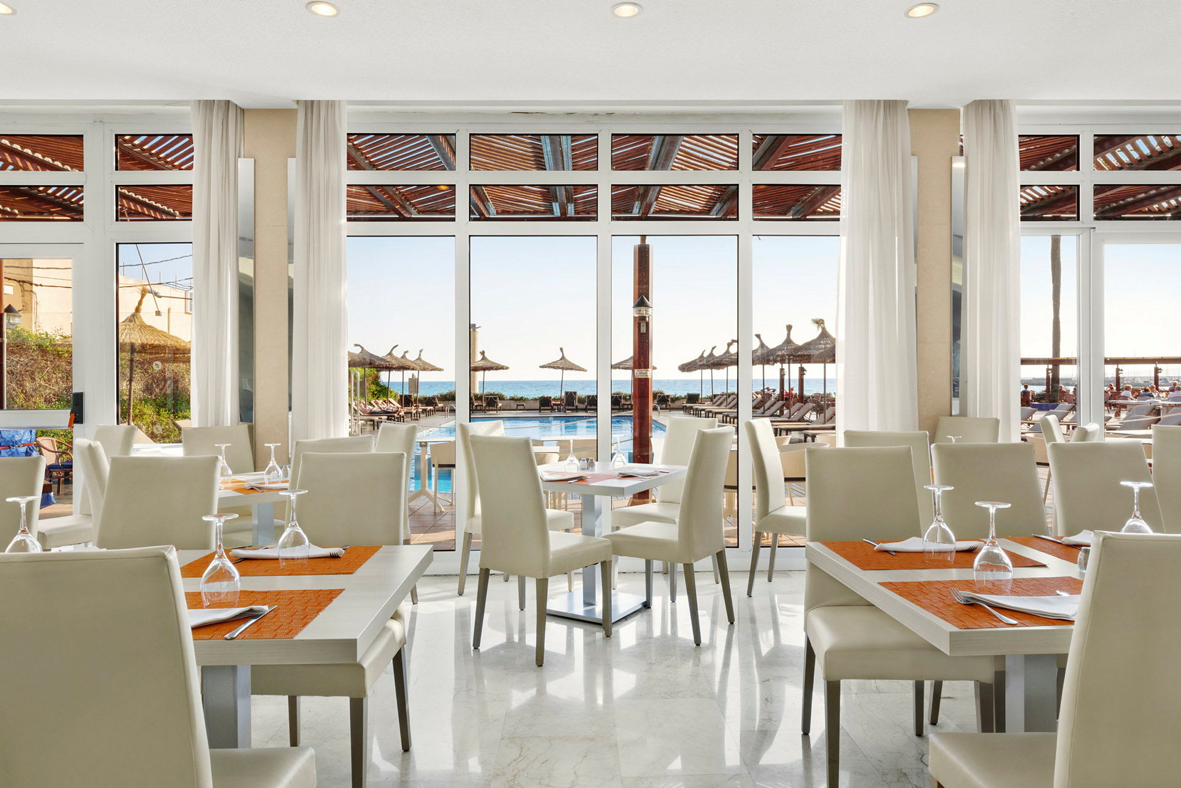 En restaurant med hvide stole og borde, med udsigt over en pool og havet gennem store glasvinduer. (AI-genereret tekst)