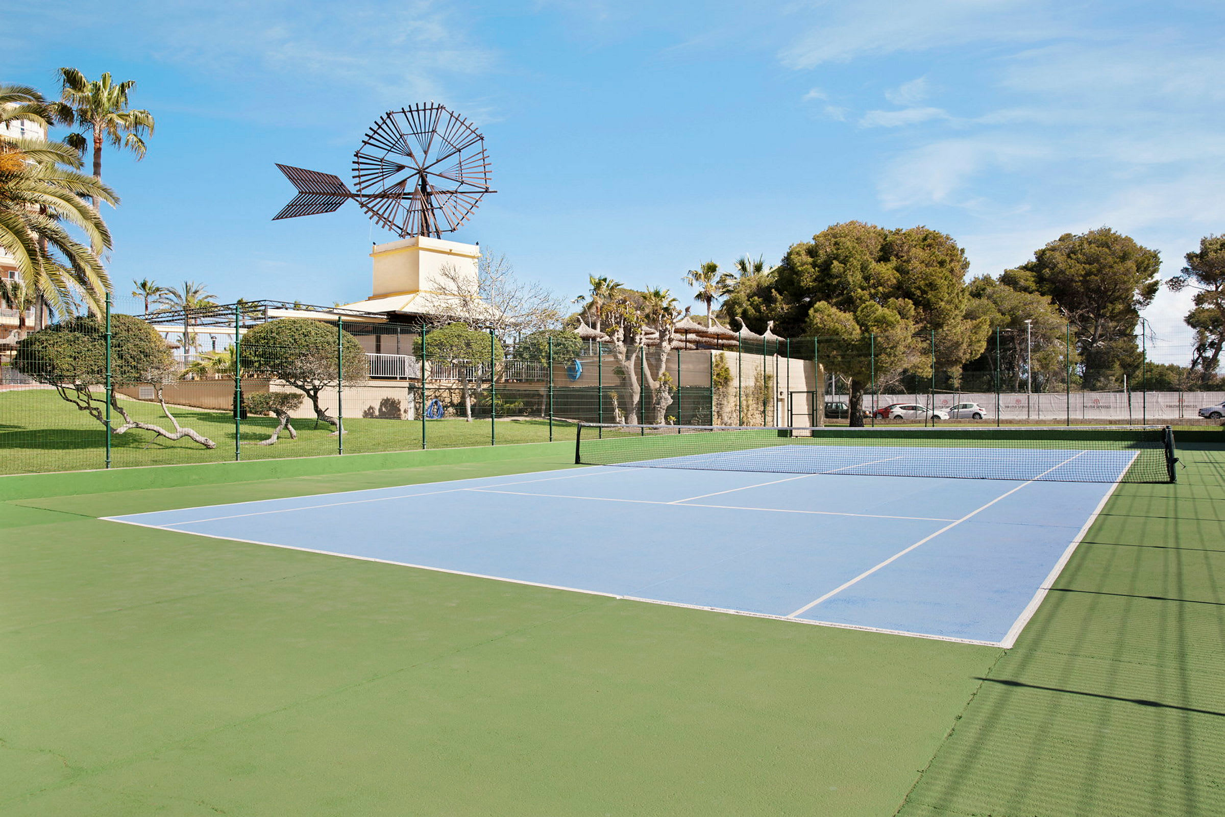 En udendørs tennisbane med en vindmølle i baggrunden og omkringliggende grønne områder. (AI-genereret tekst)