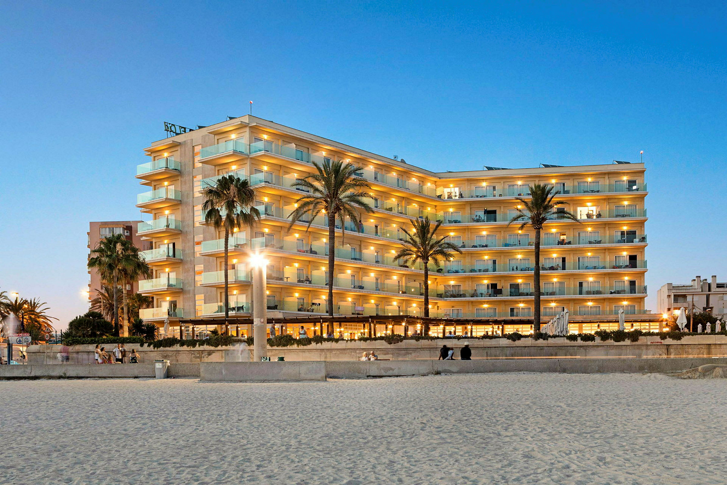 Hotelbygning med flere etager, beliggende tæt på stranden, oplyst om aftenen med palmer i forgrunden. (AI-genereret tekst)
