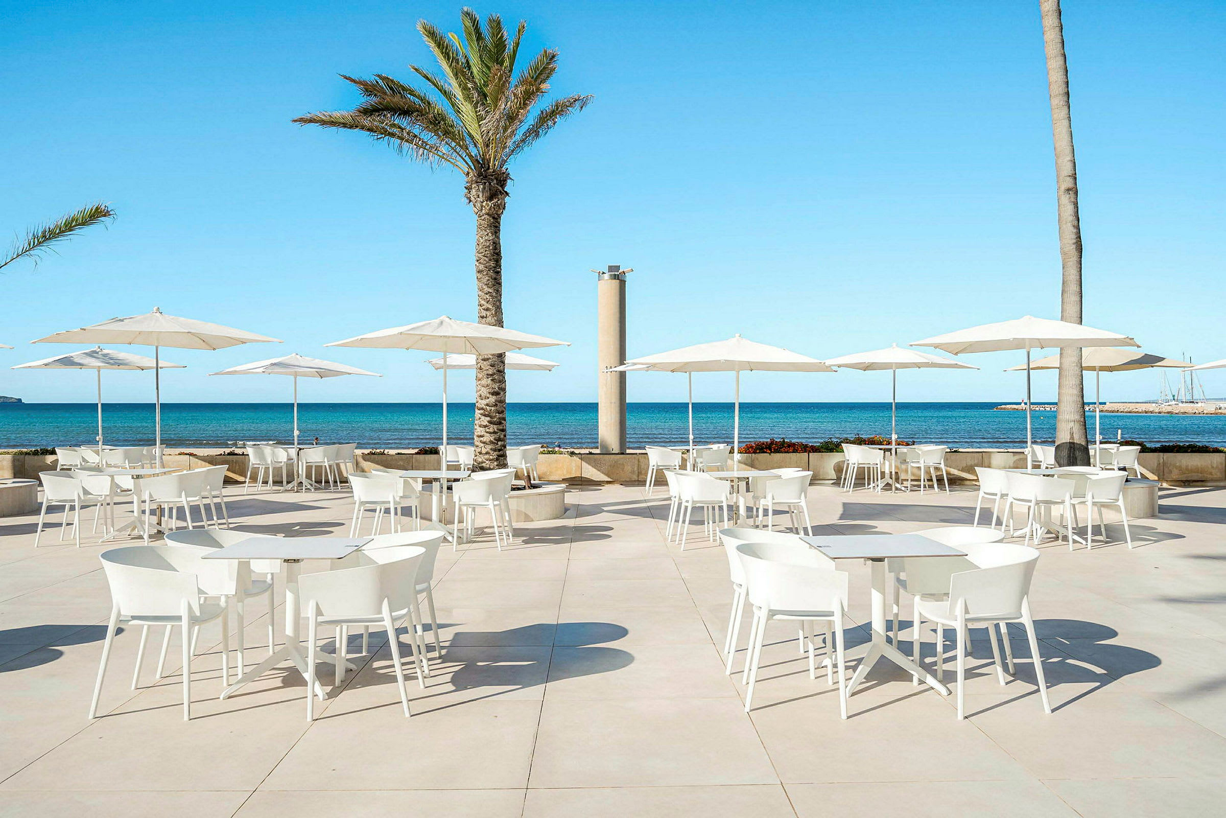 Hvidmalede borde og stole med parasoller på en terrasse ved havet. (AI-genereret tekst)