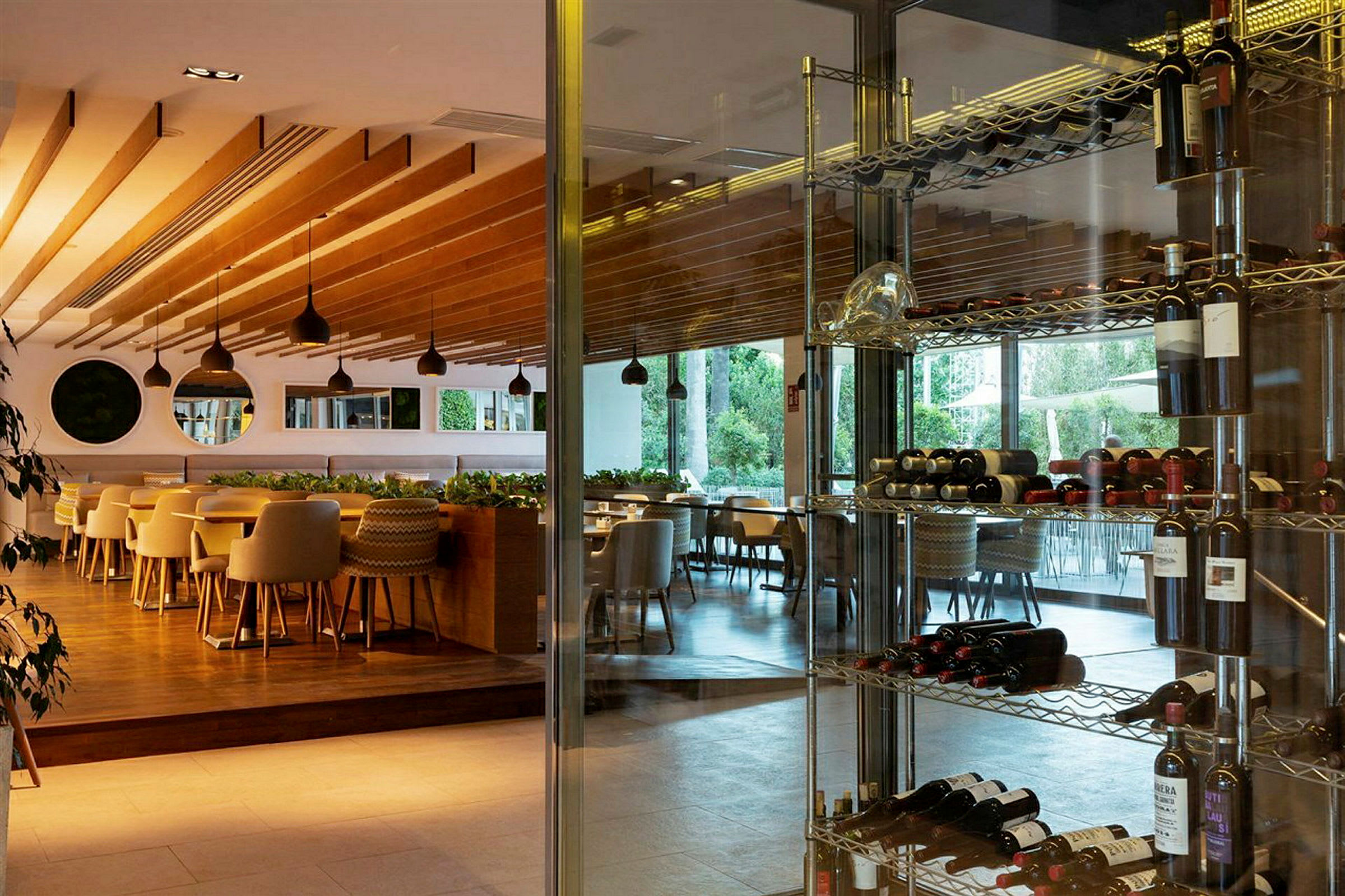 En lys restaurant med trædetaljer på loftet, store vinduer med udsigt til grønt, og vinflasker på metalhylder. (AI-genereret tekst)