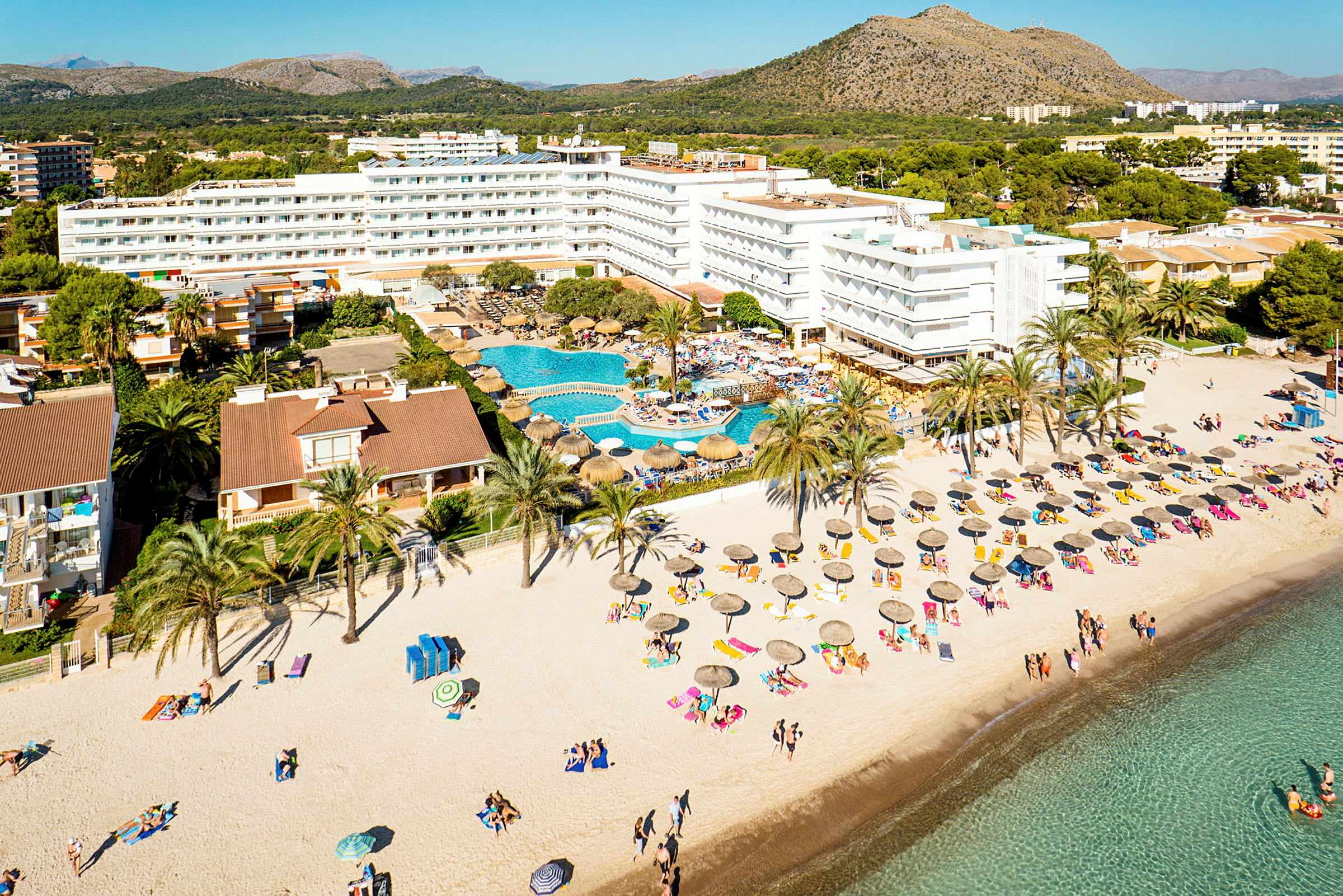 Luftfoto af et strandhotel med pool og parasoller på sandet. (AI-genereret tekst)