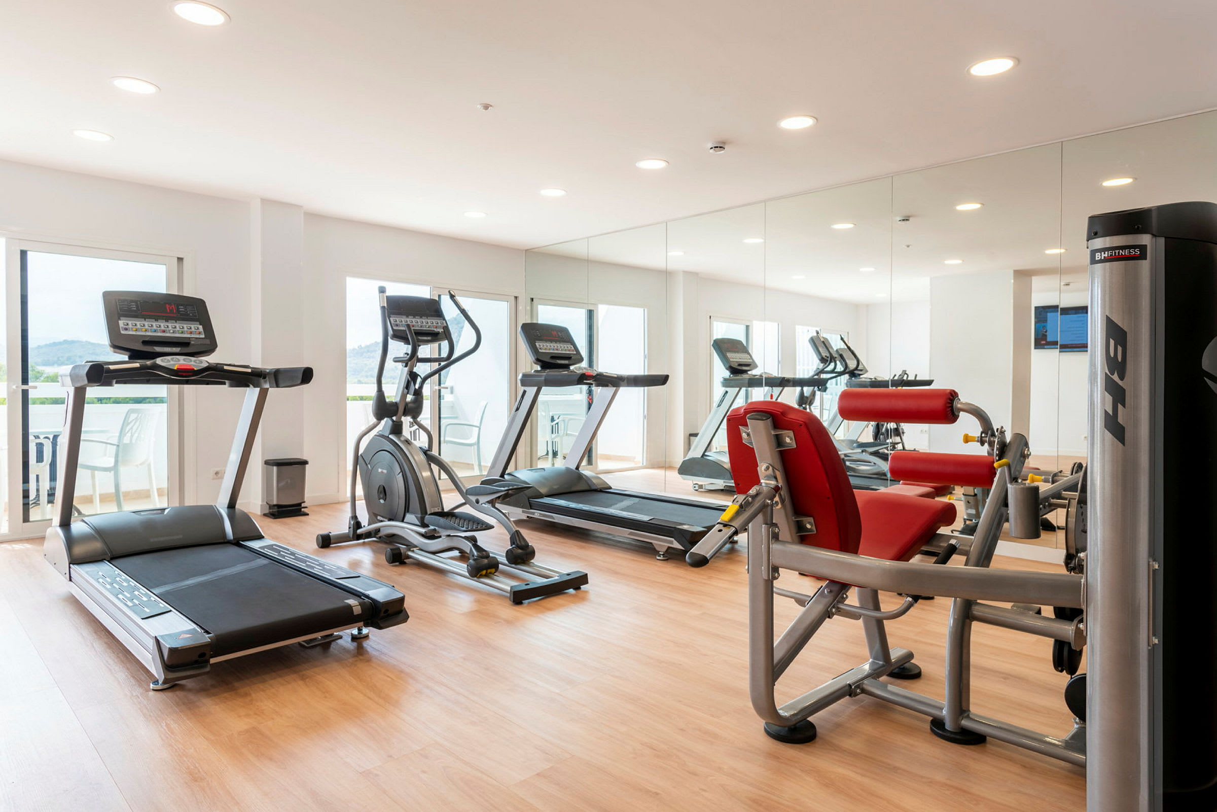 Fitnessrum med træningsmaskiner, spejlvæg og parketgulv. (AI-genereret tekst)
