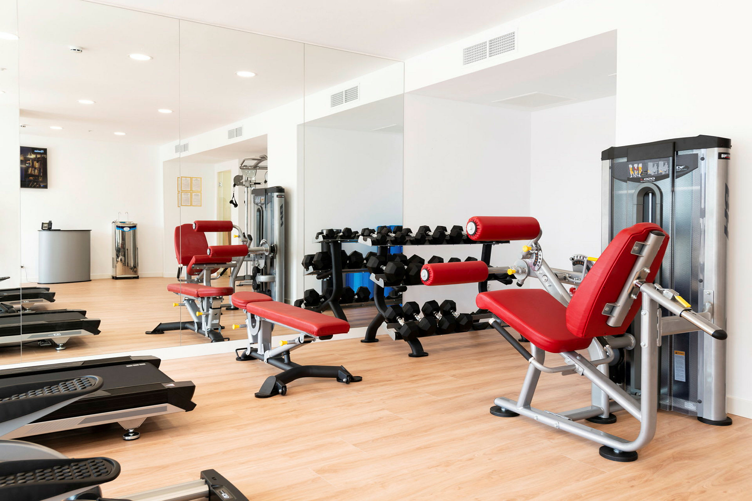 Billede af et fitnesscenter med træningsudstyr, rød bænk og spejlvæg. (AI-genereret tekst)