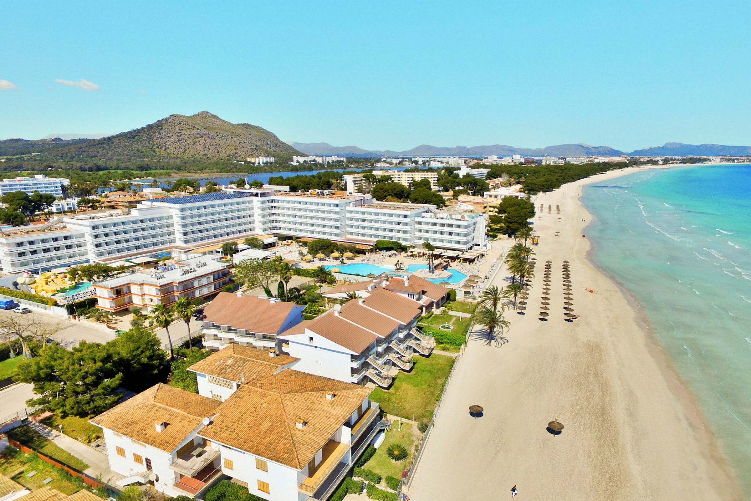 Hotelbygninger og poolområde ved en lang sandstrand med parasoller samt bjerge og vegetation i baggrunden. (AI-genereret tekst)