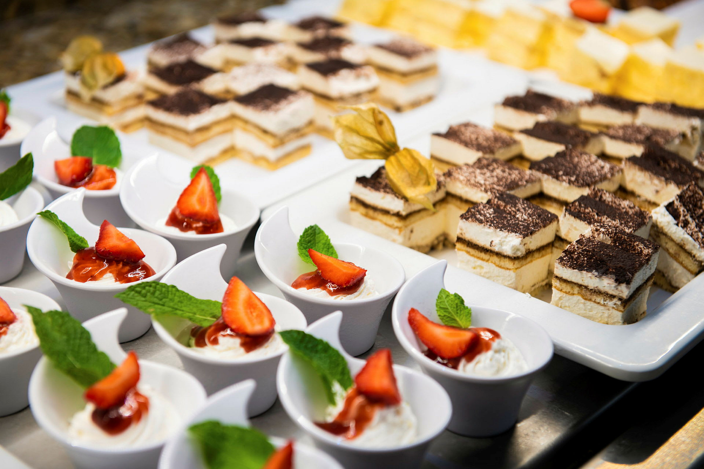 En buffet med individuelle desserter, herunder hvide skåle med fløde, jordbær og mynte samt firkantede kager med glasur. (AI-genereret tekst)