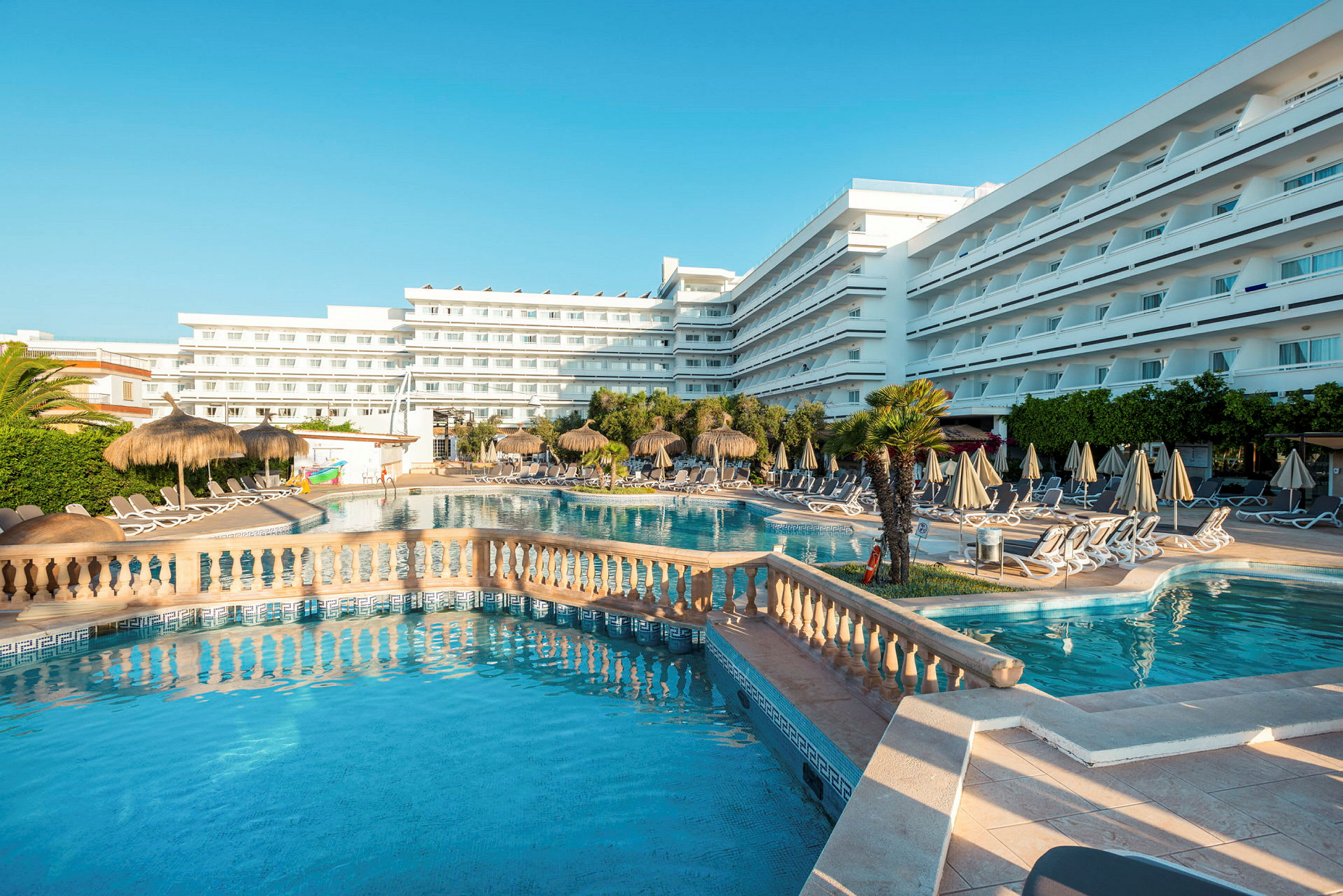 Stor hotelpool omgivet af liggestole, parasoller og grønne planter mod en baggrund af hvide bygninger. (AI-genereret tekst)