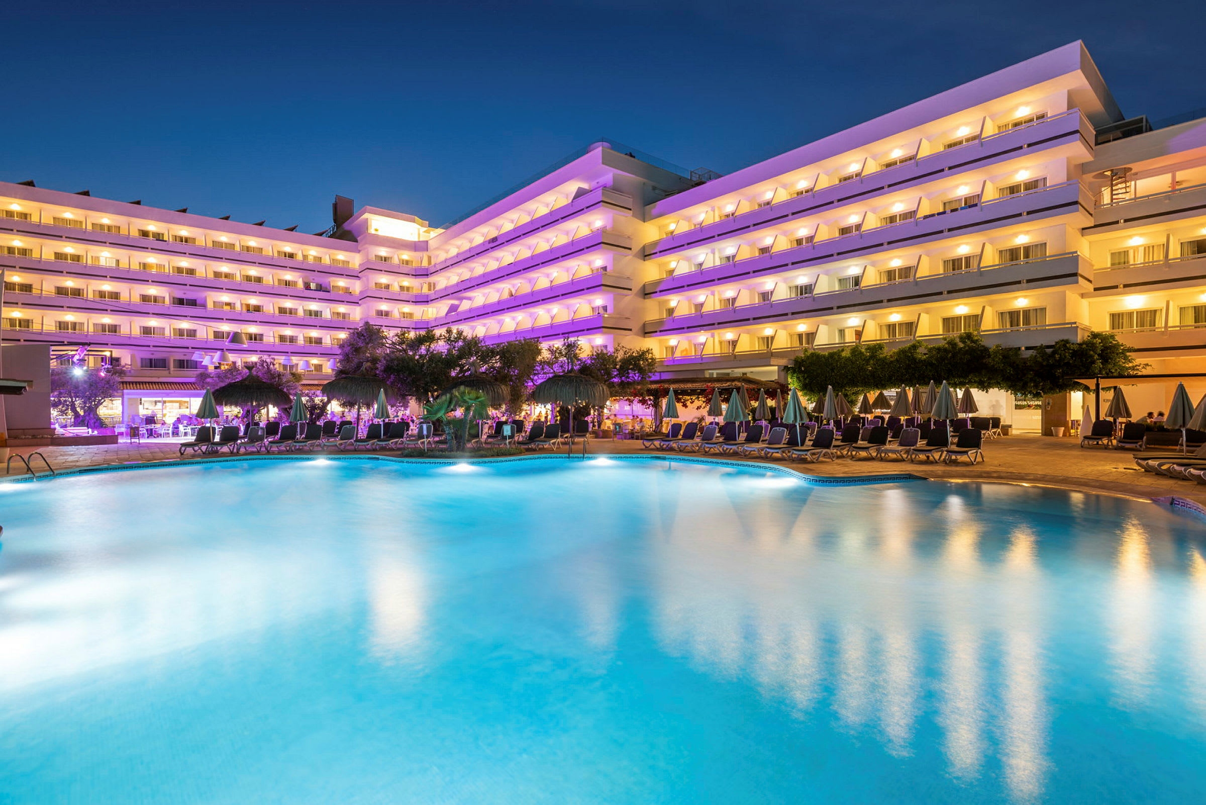 Moderne hotelbygninger med oplyste altaner synlige ved en stor pool omgivet af liggestole og parasoller om aftenen. (AI-genereret tekst)