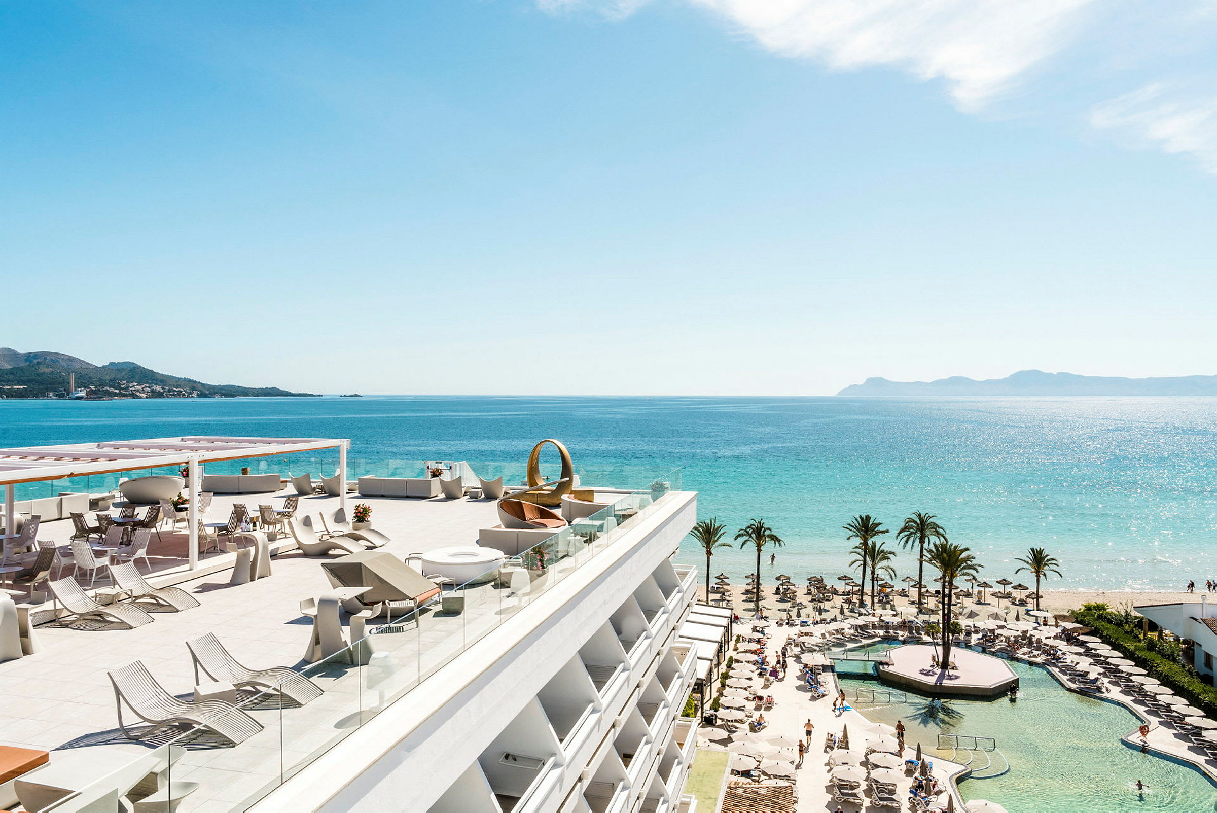 Viser en højt terrasse med liggestole og borde, kystudsigt med strand, palmer og hav. (AI-genereret tekst)