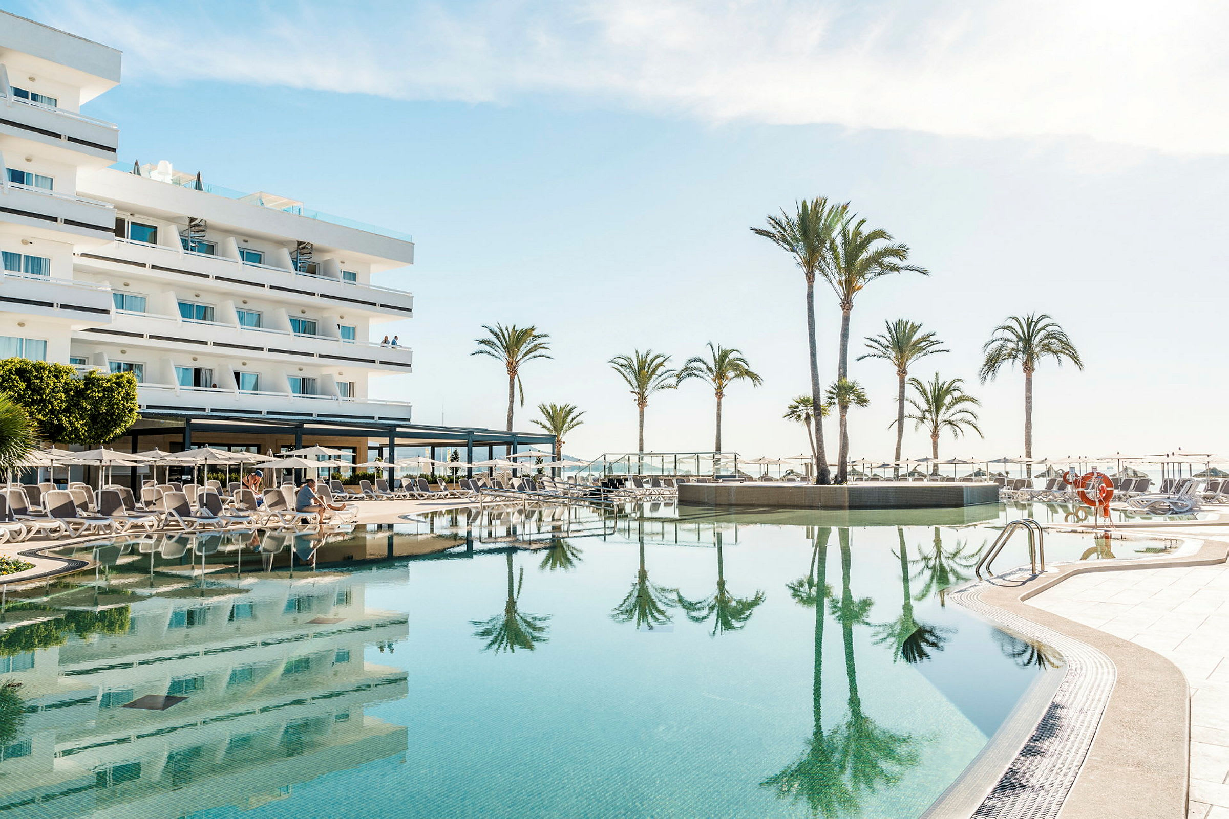 Hotel med swimmingpool og liggestole ved en solrig strand. (AI-genereret tekst)