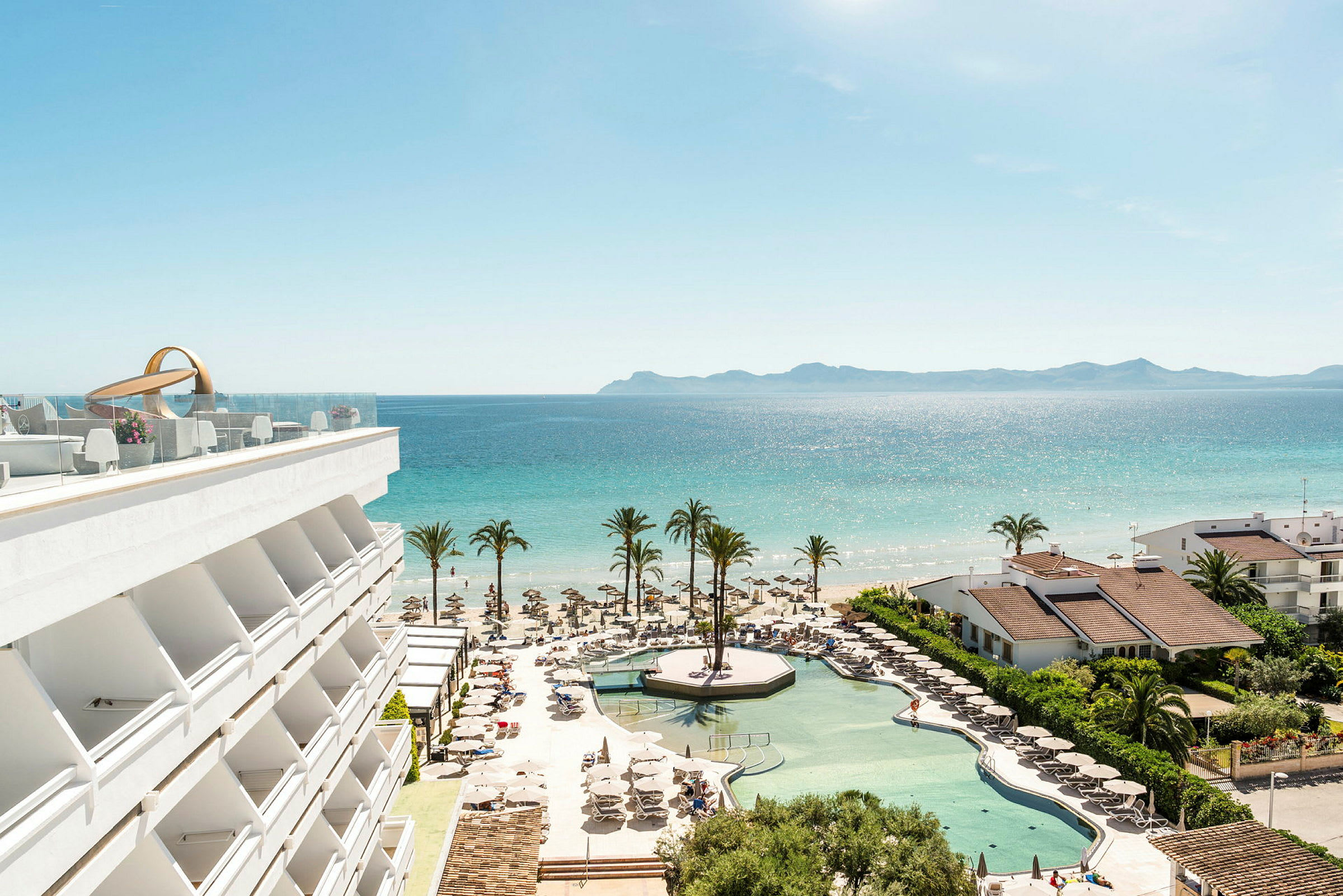 En udsigt over hotelpool, strand og hav med palmer og bygninger. (AI-genereret tekst)