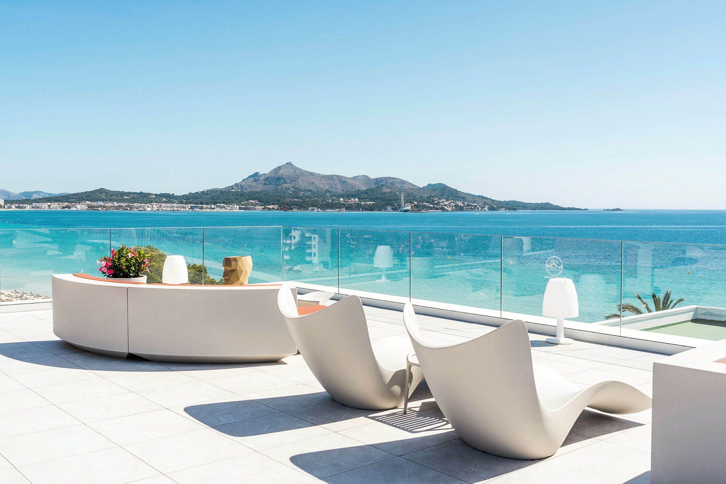 Udsigt fra balkon med moderne siddemøbler og glasrækværk mod havet og en bjergkystlinje. (AI-genereret tekst)