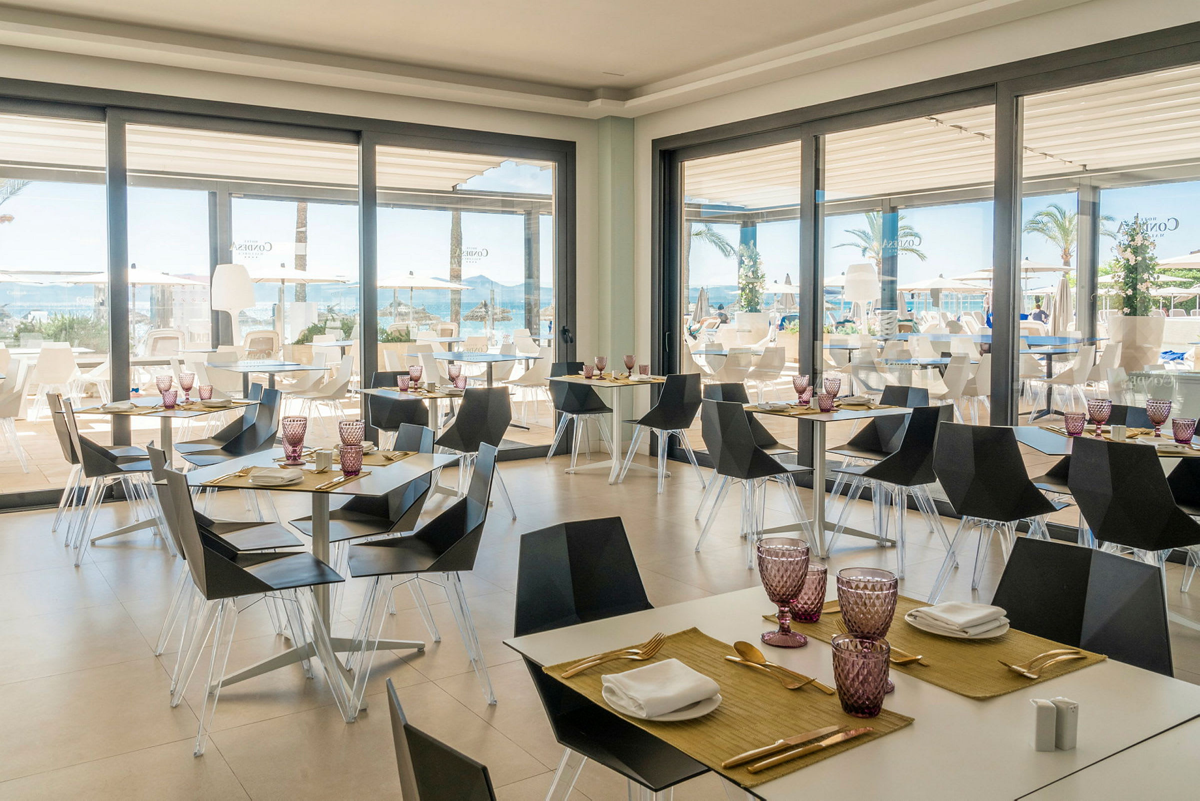 Restaurant med moderne møbler og dækkede borde ved store glasvinduer med udsigt til pool og hav. (AI-genereret tekst)