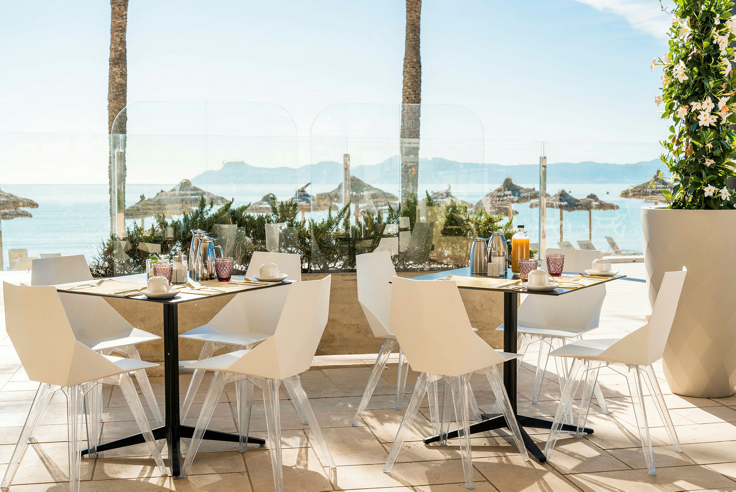 Billede af en udendørs restaurant med hvide stole og borde, med havudsigt og parasoller i baggrunden. (AI-genereret tekst)