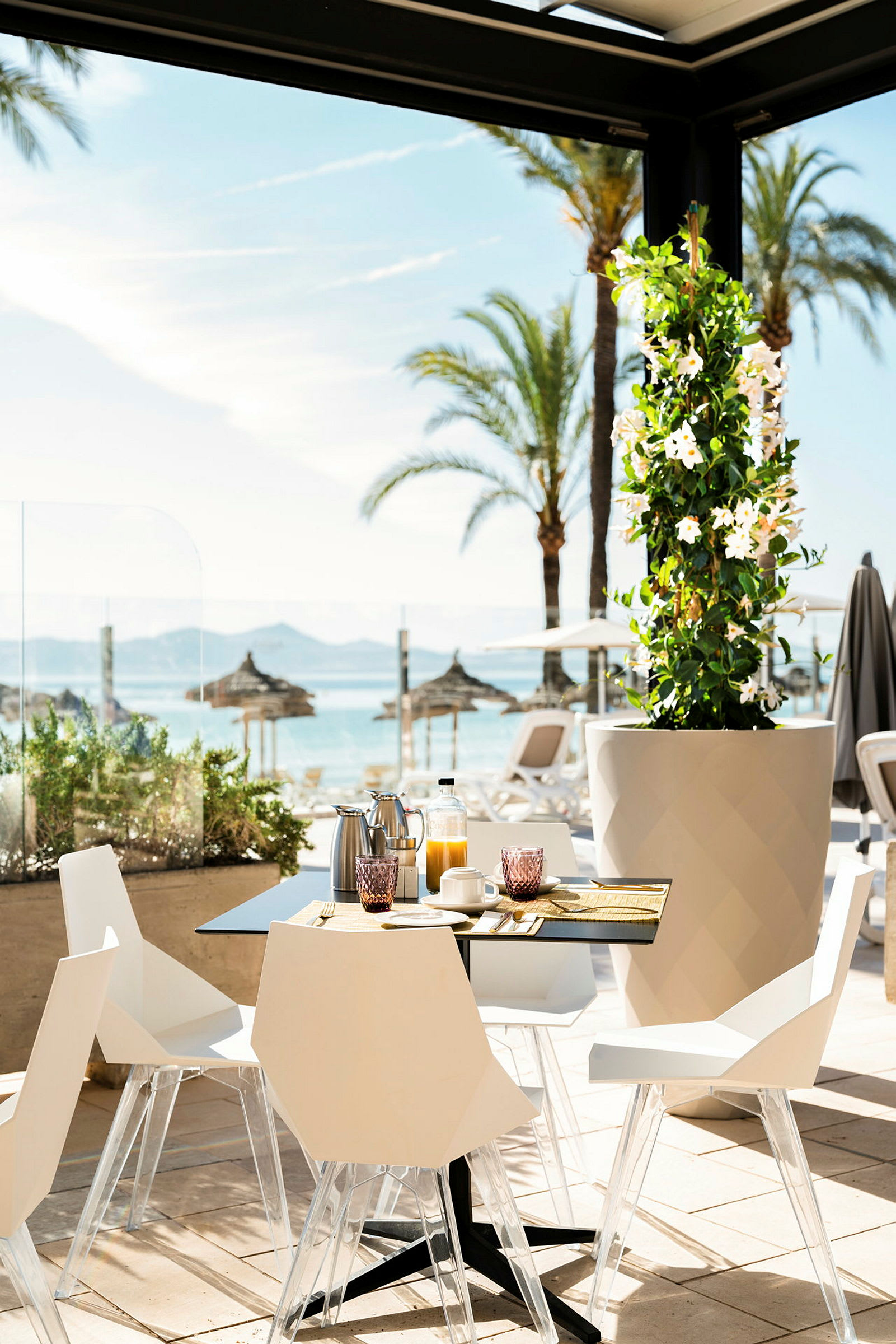 Et bord med stole, indrett til morgenmad, beliggende på en solrig terrasse med udsigt over palmer og stranden. (AI-genereret tekst)