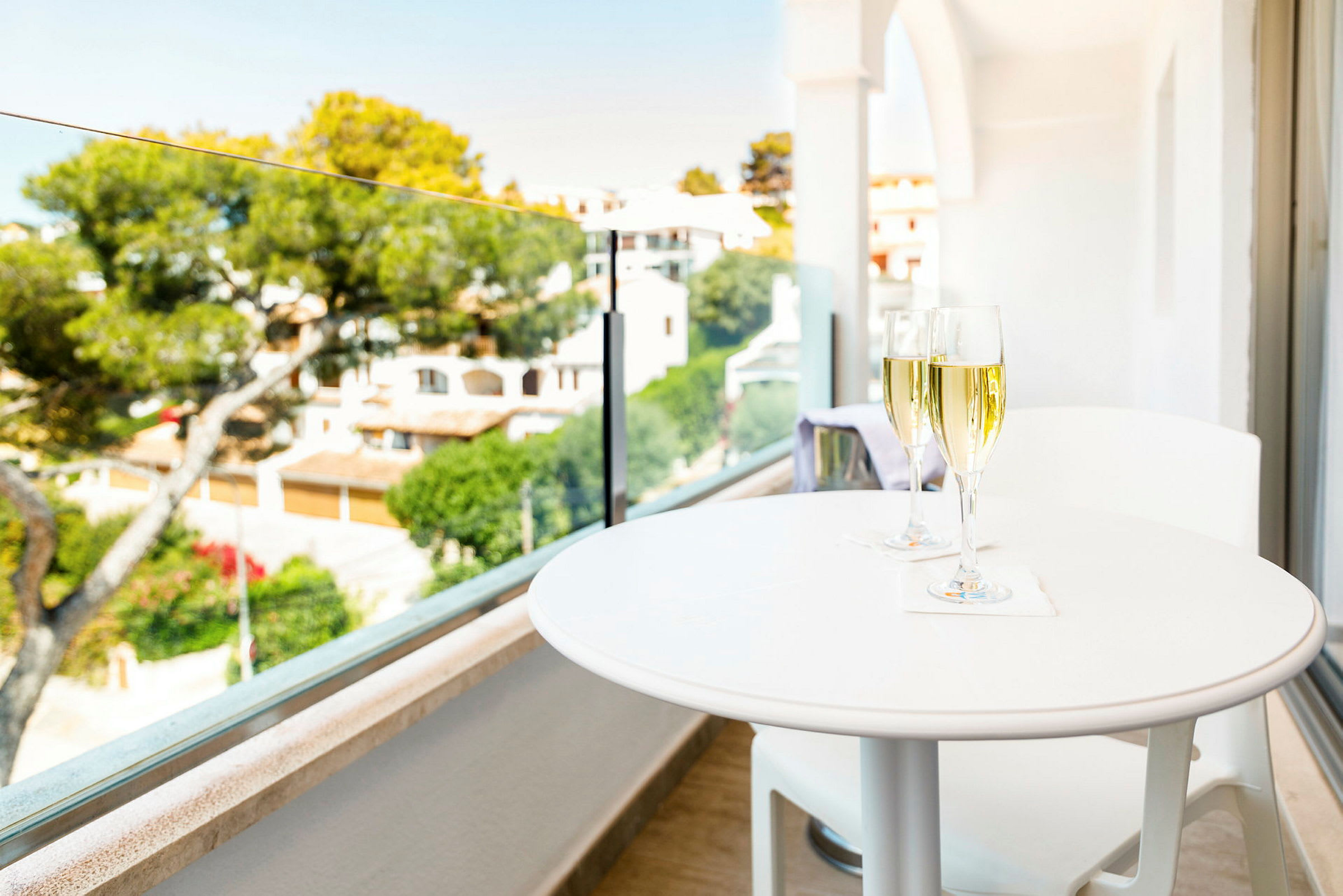 Et hvidt bord med to glas champagne på en altan med udsigt over en frodig have og huse i baggrunden. (AI-genereret tekst)