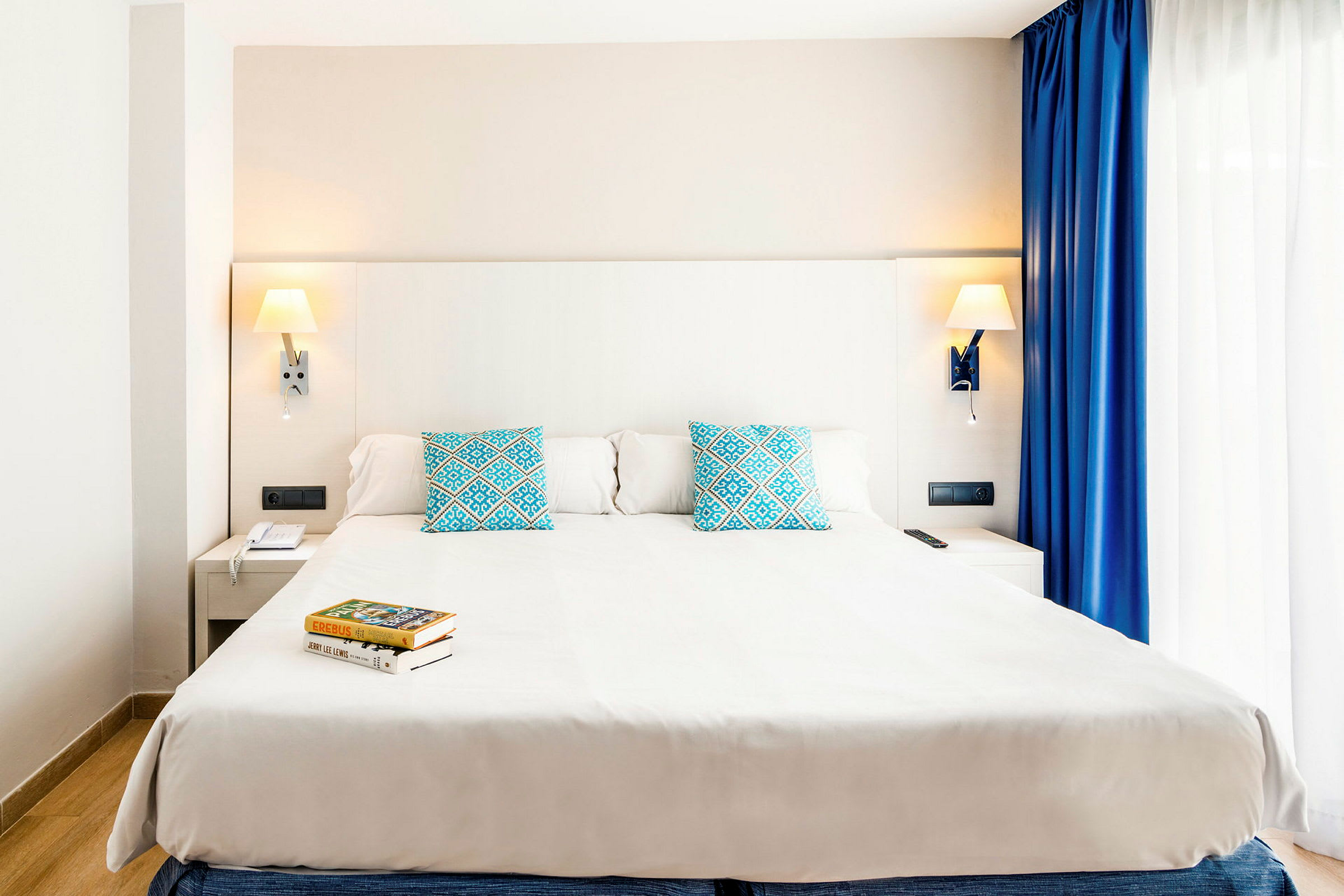 Et moderne hotelværelse med en stor dobbeltseng, to blå pyntepuder med mønster og vægmonterede læselamper. Ved siden af sengen er der et natbord og et vindue med hvide og blå gardiner. (AI-genereret tekst)
