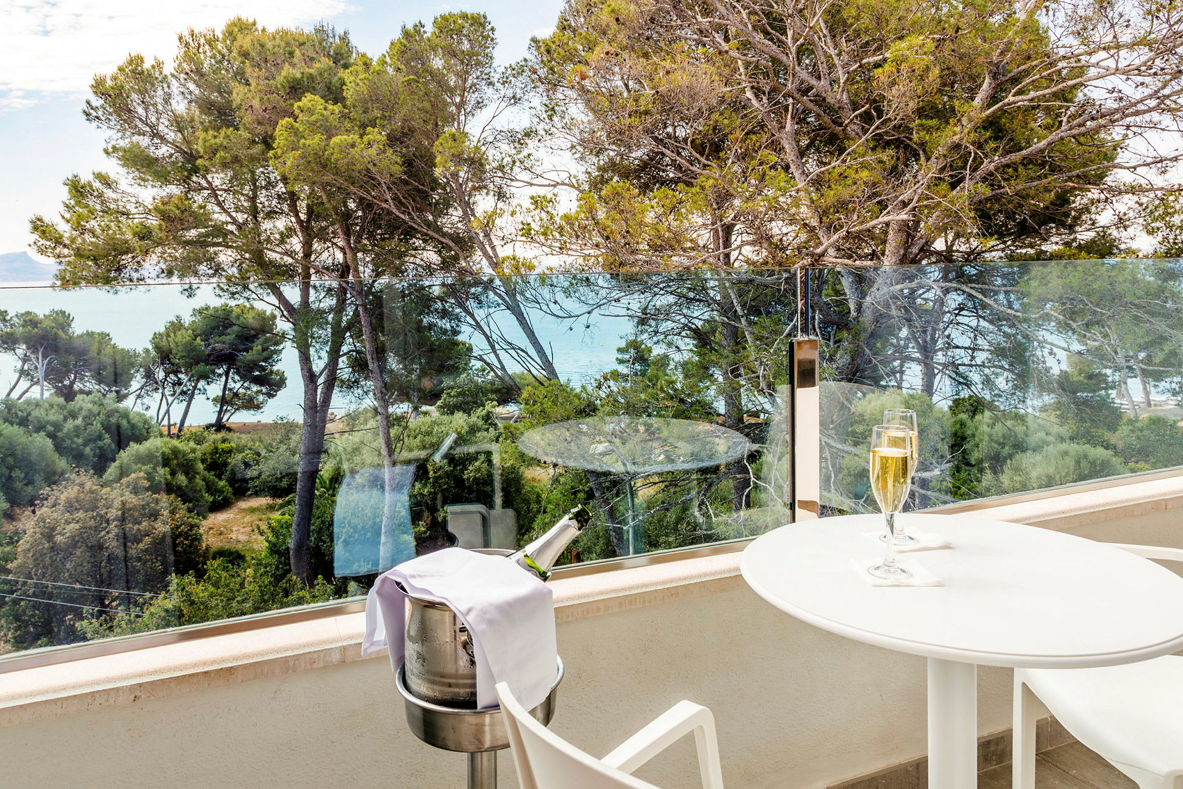 En balkon med udsigt over træer og havet. På balkonen står et hvidt bord med et glas champagne og en holder til en champagneflaske. (AI-genereret tekst)