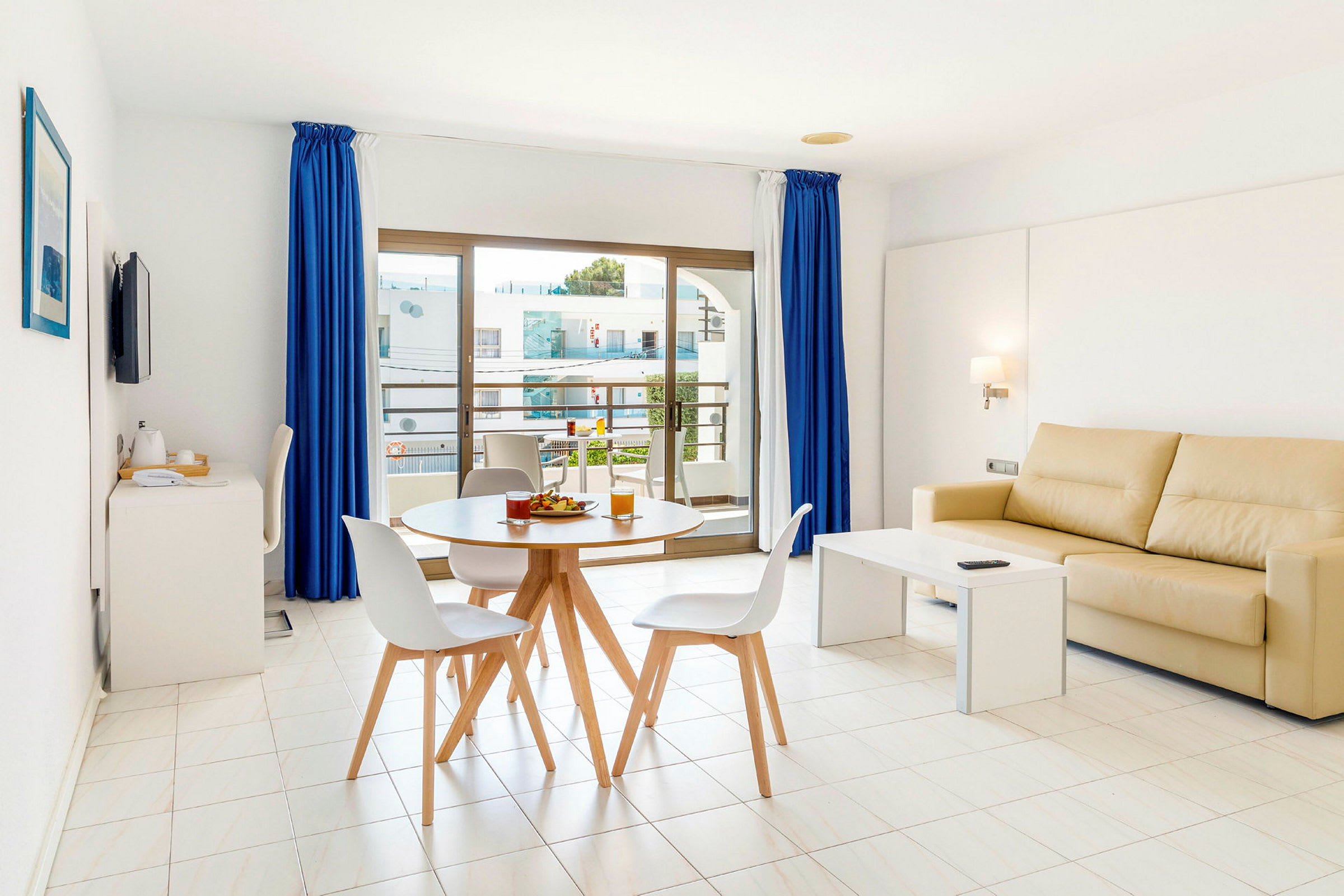 En moderne stue med lys indretning. En beige sofa står ved et hvidt sofabord, og et rundt spisebord med stole er placeret foran store vinduer med blå gardiner. (AI-genereret tekst)