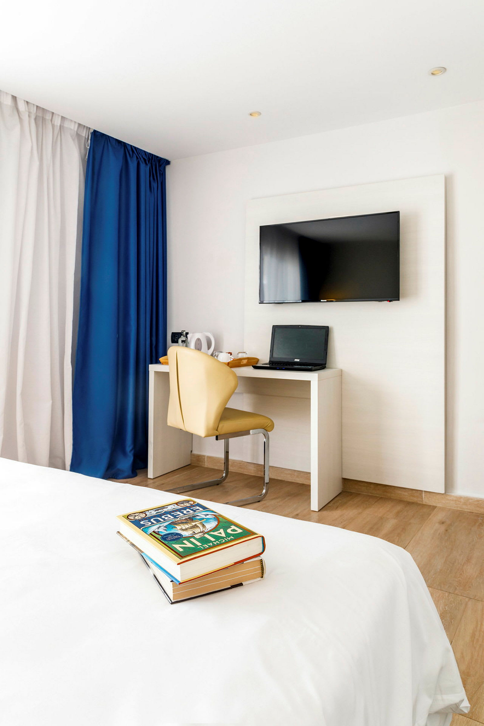 Et hotelværelse med et fladskærms-tv på væggen, et skrivebord med en bærbar computer og en beige stol. I forgrunden ses en seng med en stak bøger. (AI-genereret tekst)