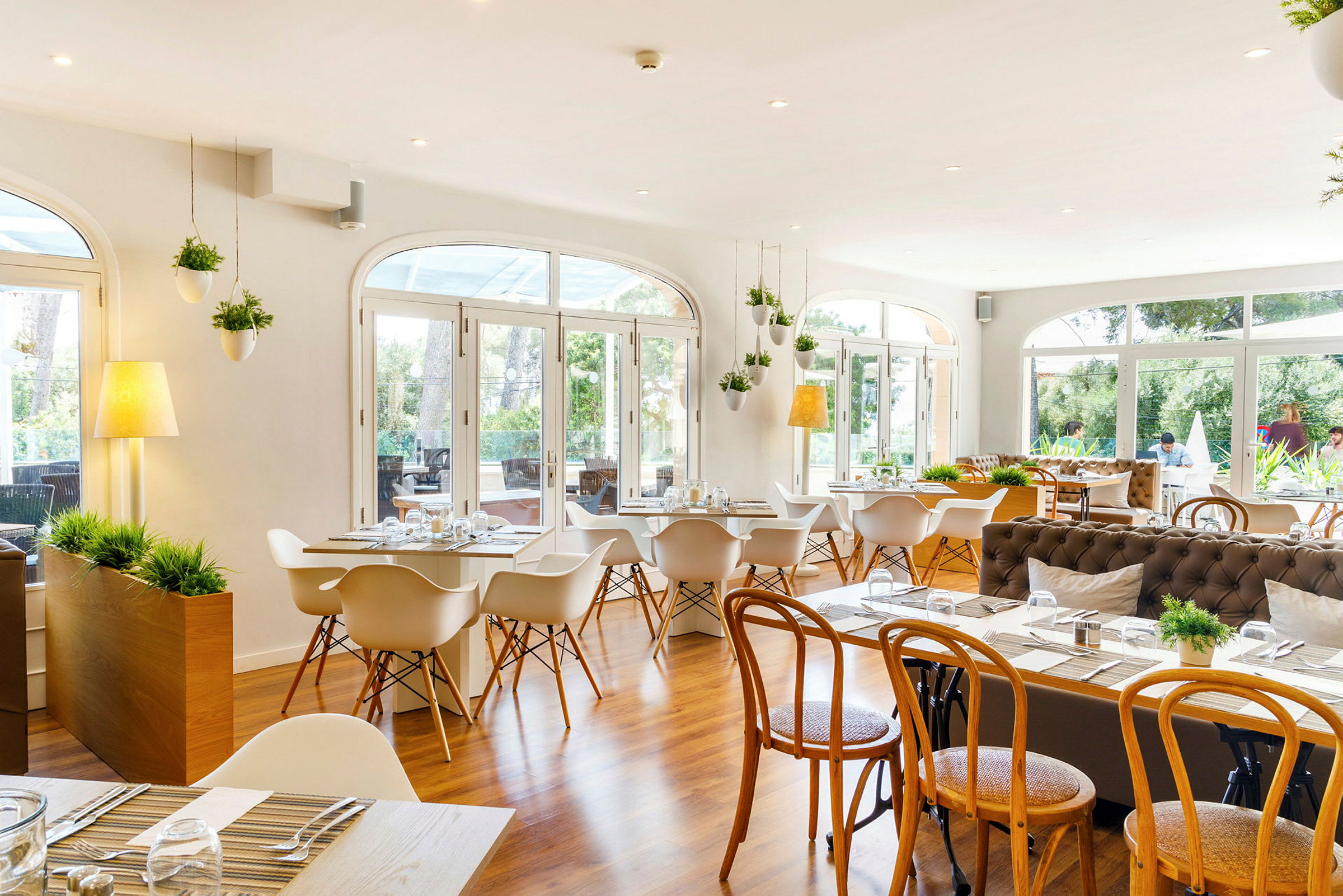 Et lyst og rummeligt restaurantområde med store vinduer, der vender ud mod en grøn udsigt. Rummet er indrettet med moderne hvide stole og træmøbler, med dekorative planter, der hænger fra loftet. (AI-genereret tekst)