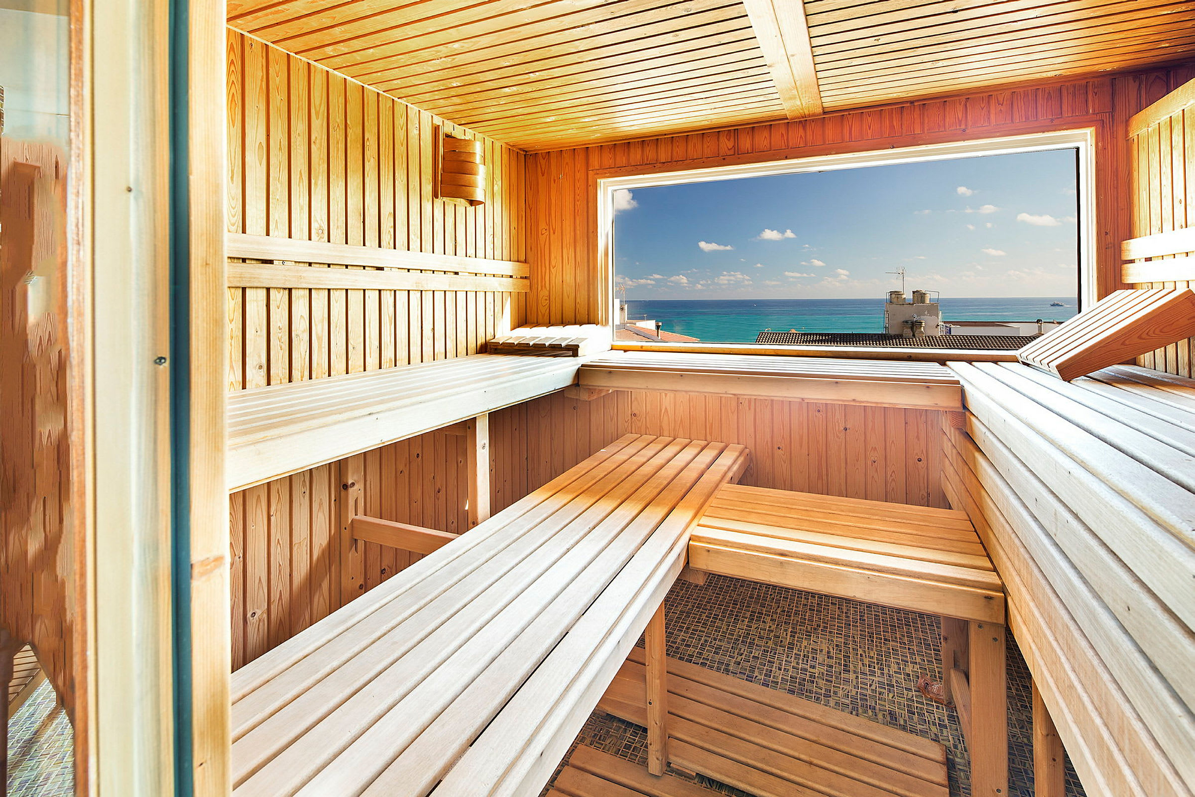 En sauna med træbænke og et stort vindue, der vender ud mod havet med klart blåt vand. (AI-genereret tekst)