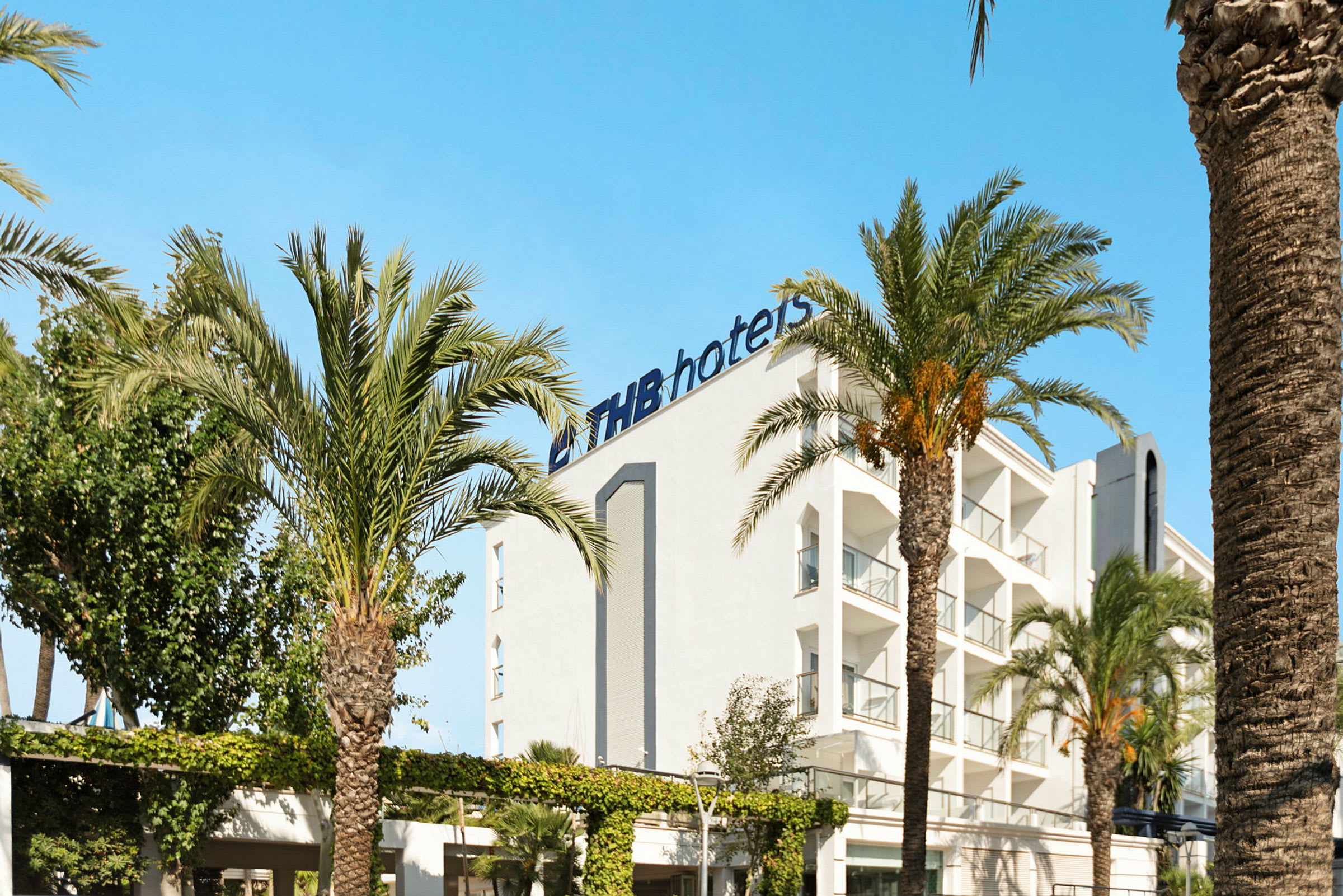 Hvid bygning med logoen 'THB hotels' omgivet af palmer under en klar blå himmel. (AI-genereret tekst)