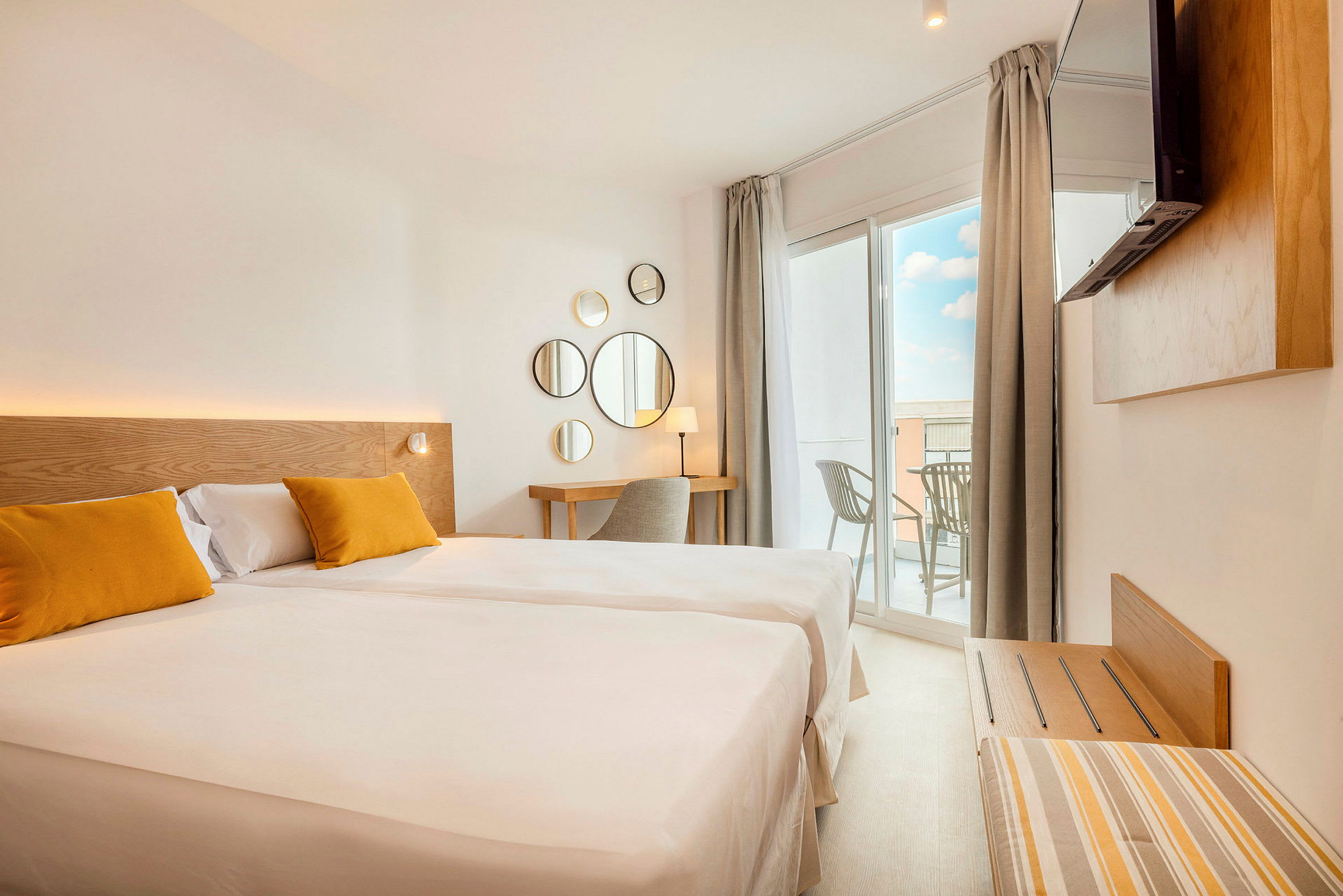 Et lyst hotelværelse med en stor seng, to gule puder, et skrivebordshjørne og en balkon med udsigt. (AI-genereret tekst)