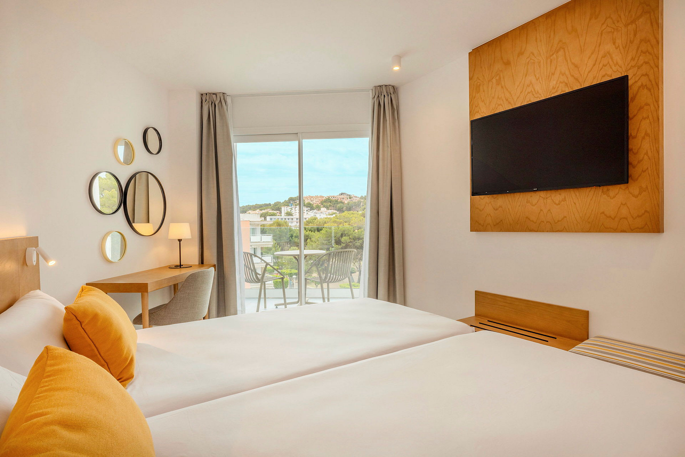 Et moderne hotelværelse med to senge, gule puder, et tv monteret på væggen, et skrivebord med lampe og en altan med udsigt. (AI-genereret tekst)