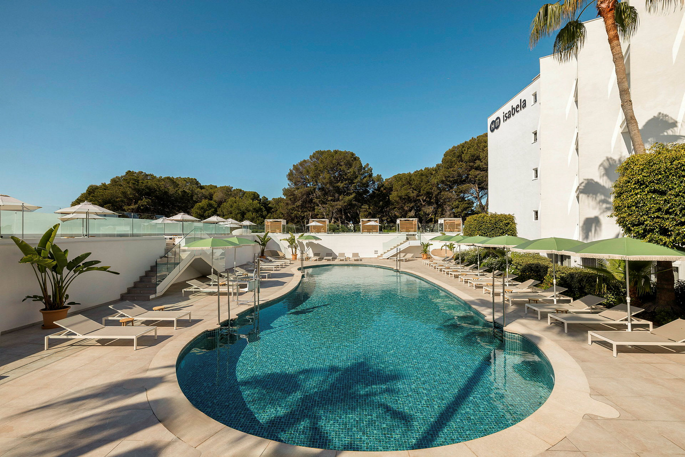 Billedet viser en udendørs pool omgivet af liggestole og parasoller, med vegetation i baggrunden og en hotelbygning til højre. (AI-genereret tekst)