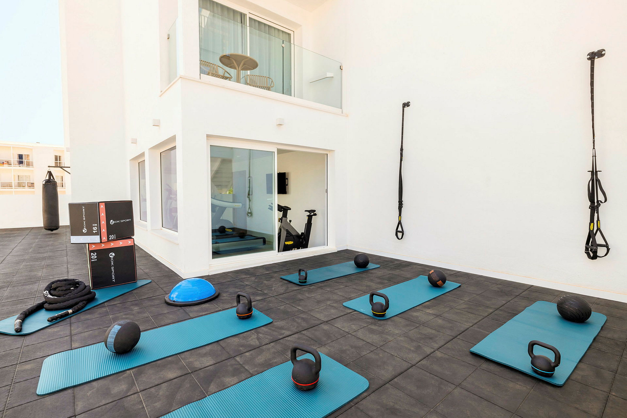 Et udendørs træningsområde med træningsmåtter, kettlebells, medicinbolde og TRX-stropper samt en boksebold. (AI-genereret tekst)