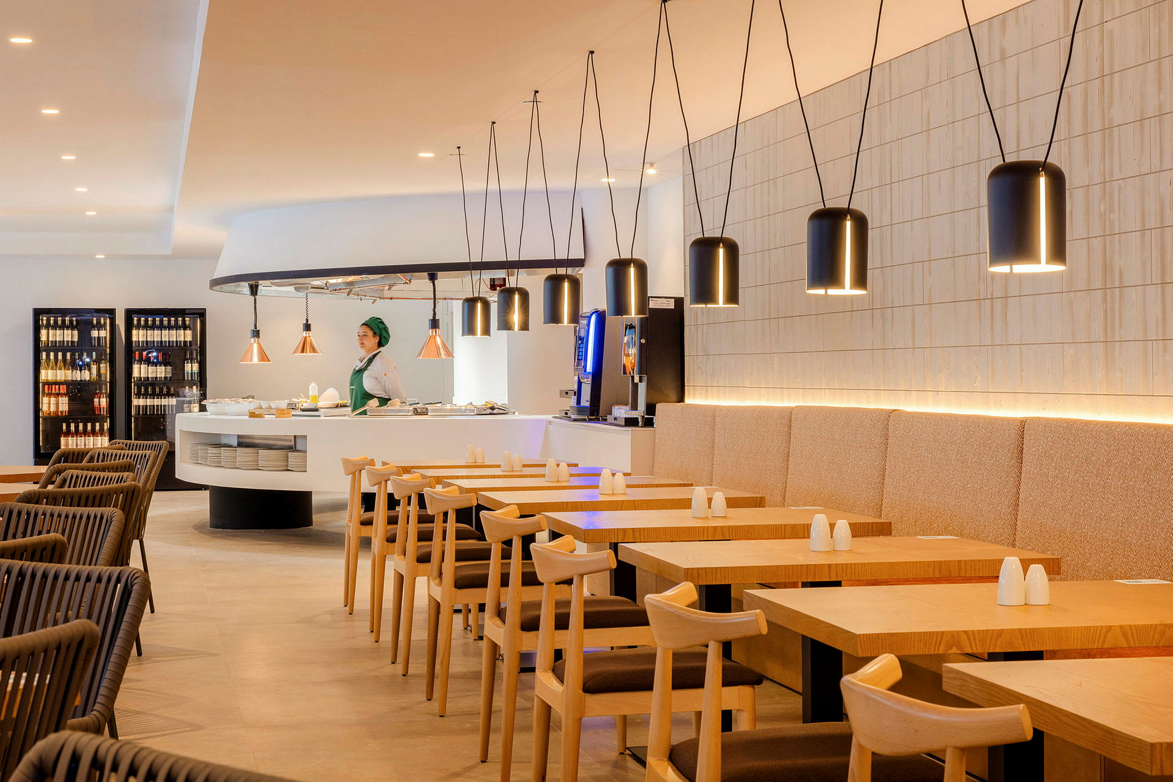Billede af en moderne restaurant med træborde og stole. En person står ved en serveringsdisk omgivet af hængende lamper. (AI-genereret tekst)