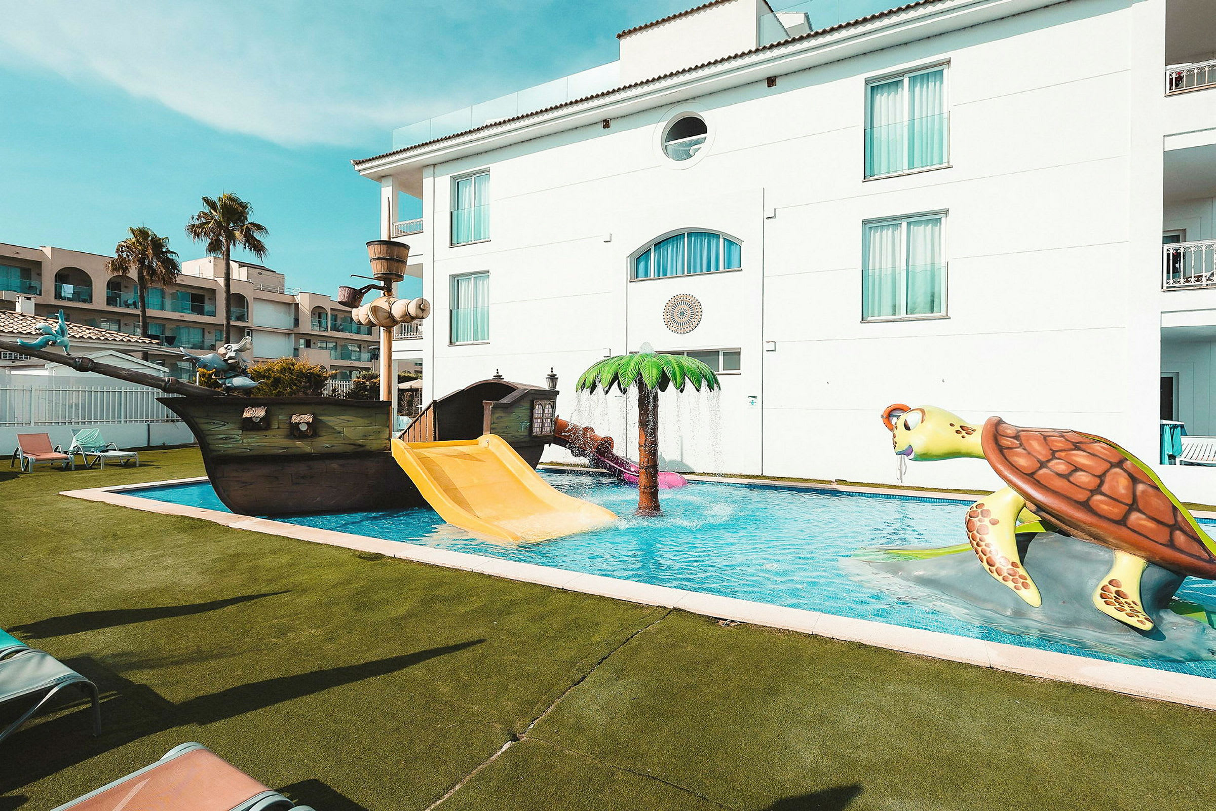 Udendørs pool med legeudstyr, herunder en skibsgynge, vandrutsjebane og en stor skildpaddefigur. (AI-genereret tekst)