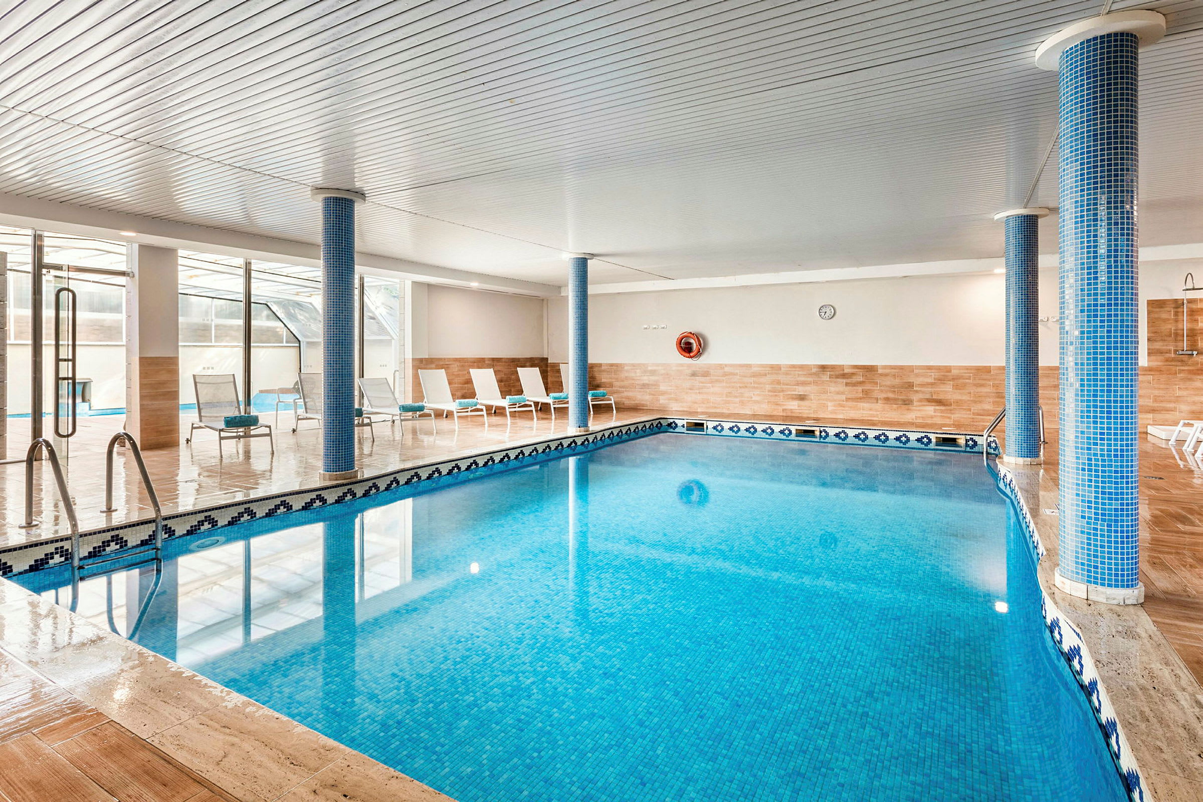 Indendørs pool med blåt vand, omgivet af solsenge og søjler. (AI-genereret tekst)