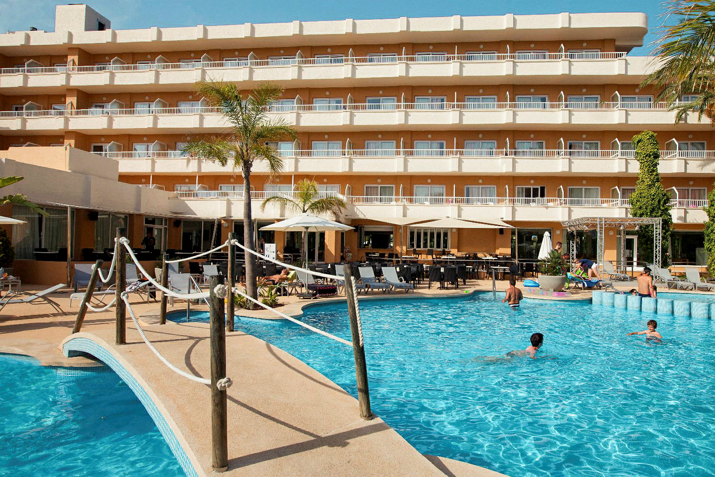 En udendørs pool foran et hotel med flere etager og altaner. (AI-genereret tekst)