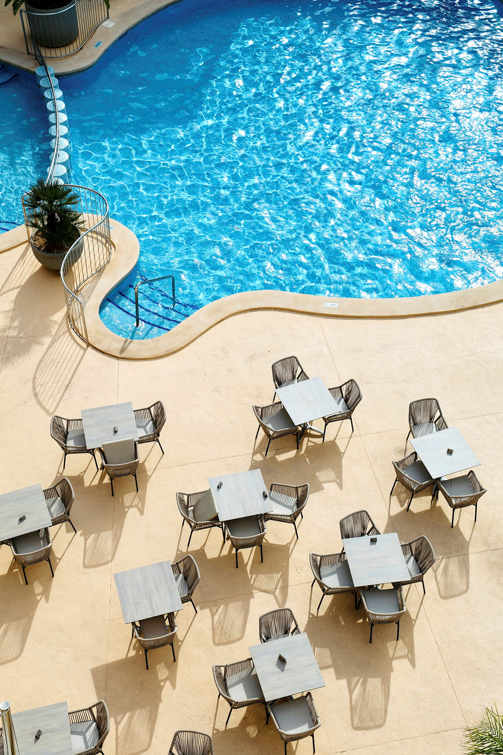 En pool med blåt vand og flere borde med stole på en solrig terrasse. (AI-genereret tekst)
