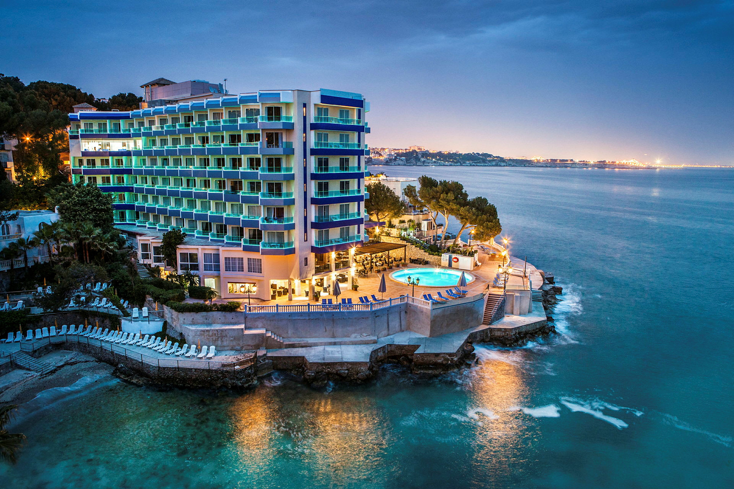 Billede af et hotel ved havet med en oplyst pool og omkringliggende bygninger. (AI-genereret tekst)